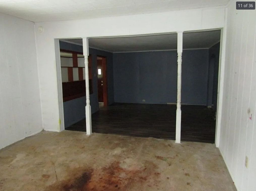 A living room with a carpet hugely stained by what looks like blood or dark red paint. It is empty and dark. 