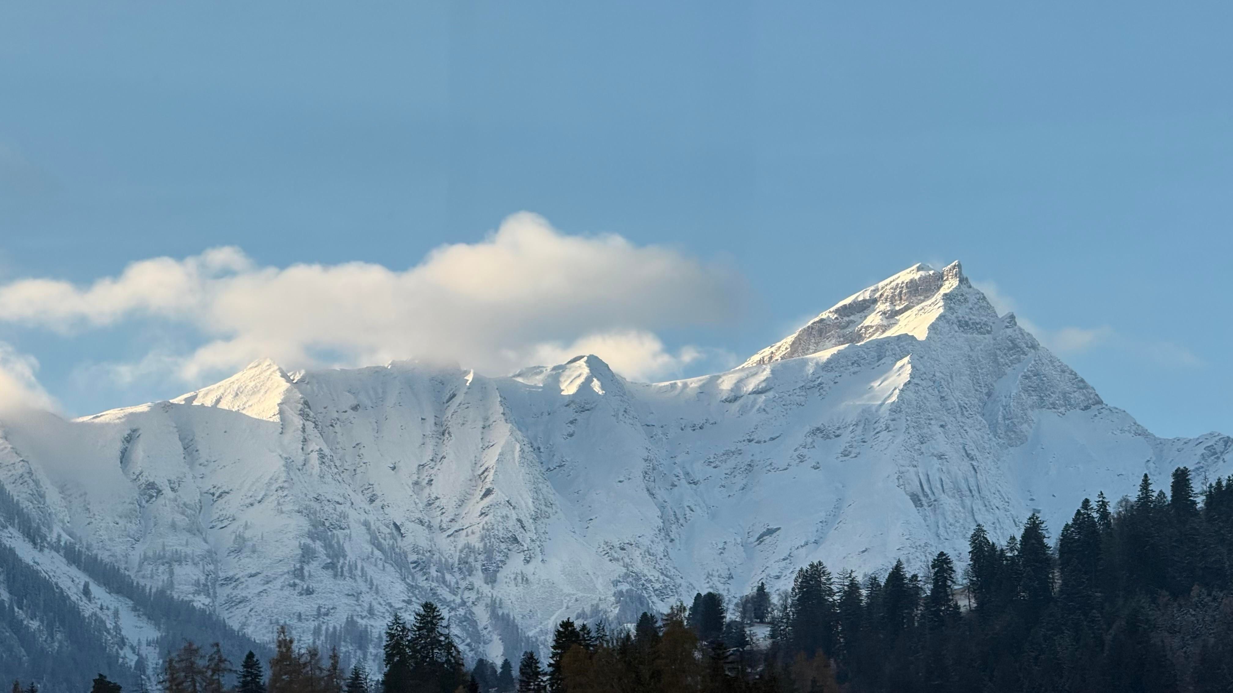 A snowy mountain range under a clear blue sky, with some clouds drifting near the peaks. Trees are visible in the foreground.