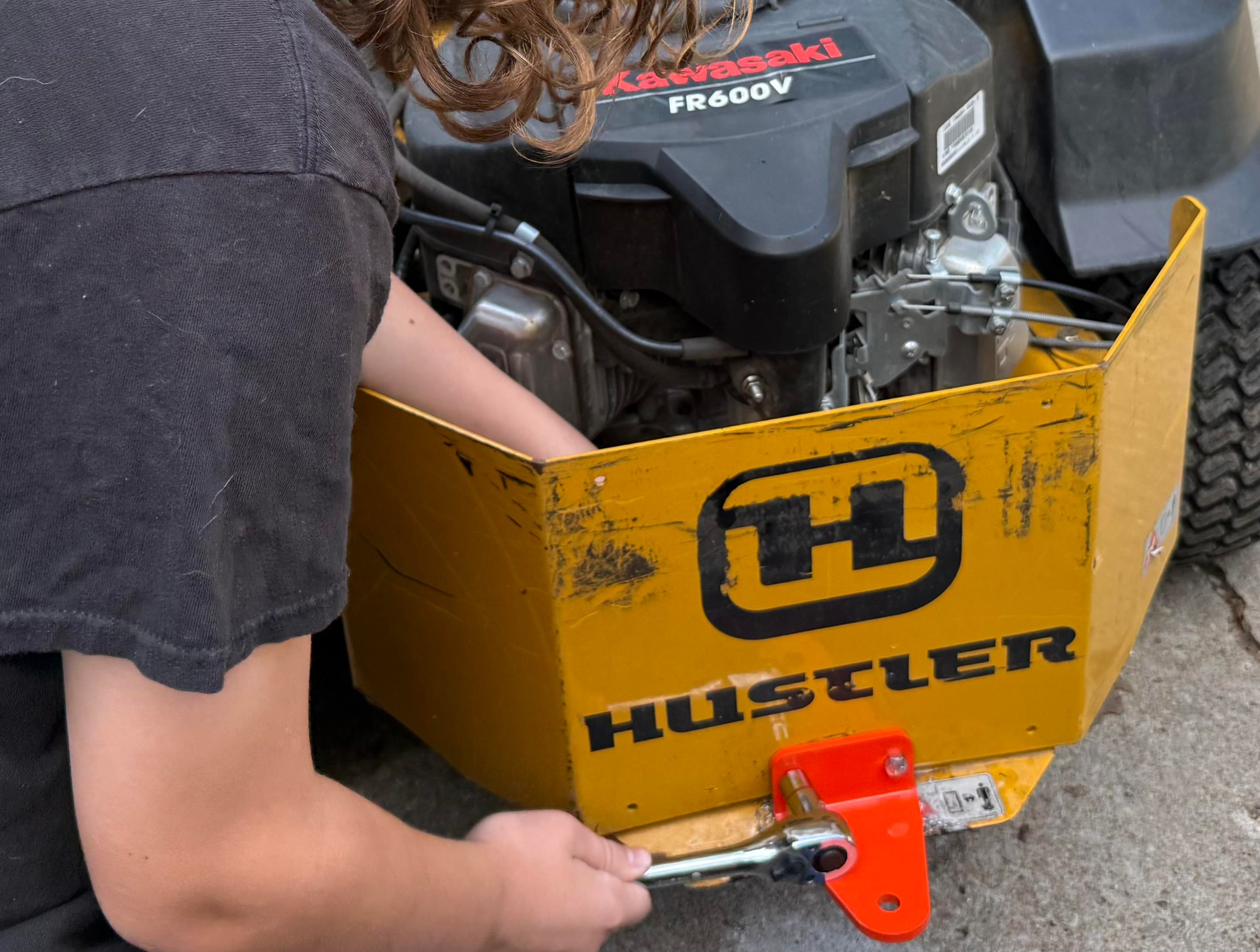Teen boy uses a socket wrench to tighten a bolt holding the new orange plate to the battle worm and scuffed yellow chassis of a hustler 