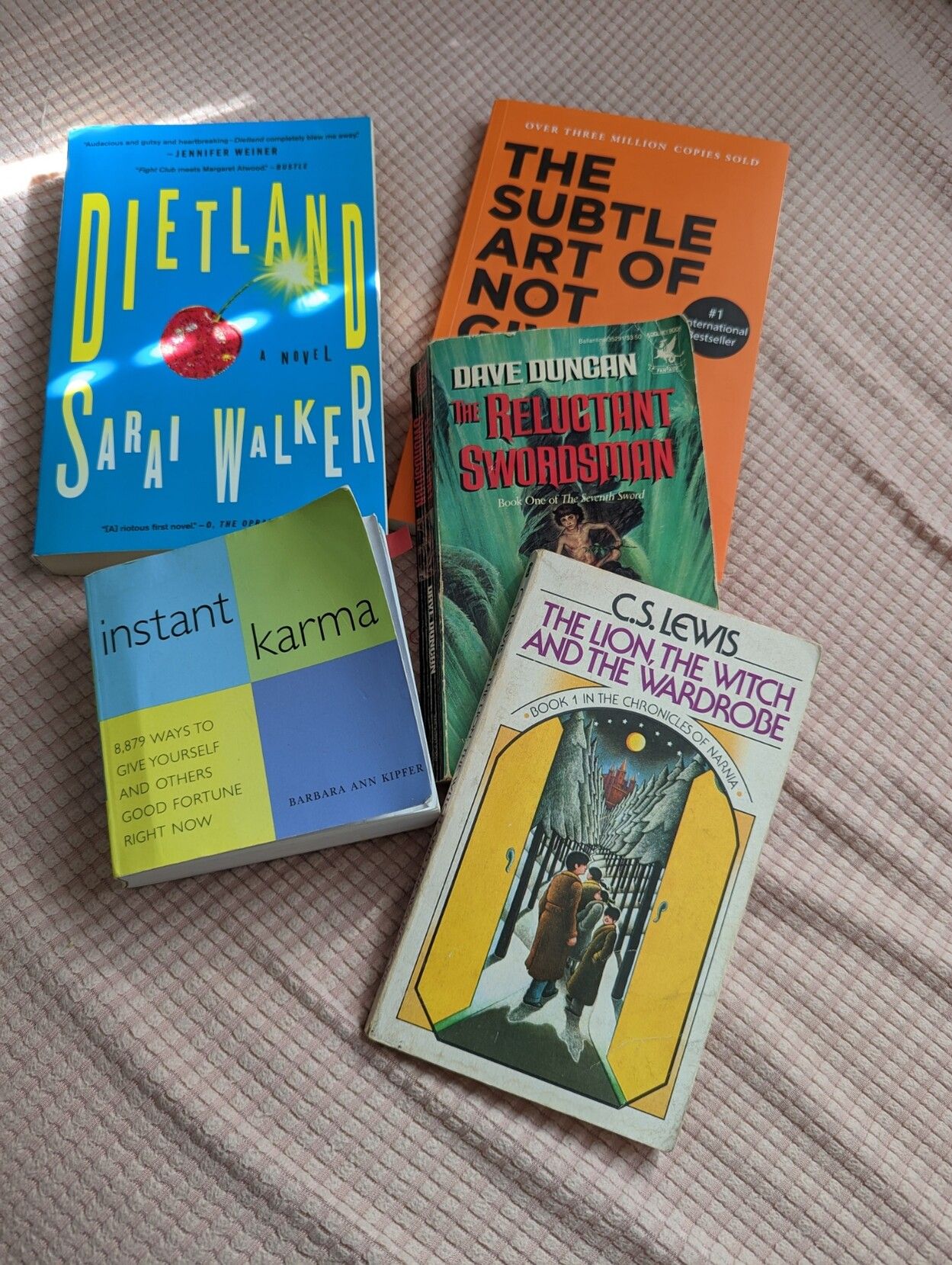 5 Books in a pile on a pink background. The titles are Dietland; The Lion, The Witch and the Wardrobe; instant karma; The Reluctant Swordsman; and The Subtle Art of Not Giving a F***.