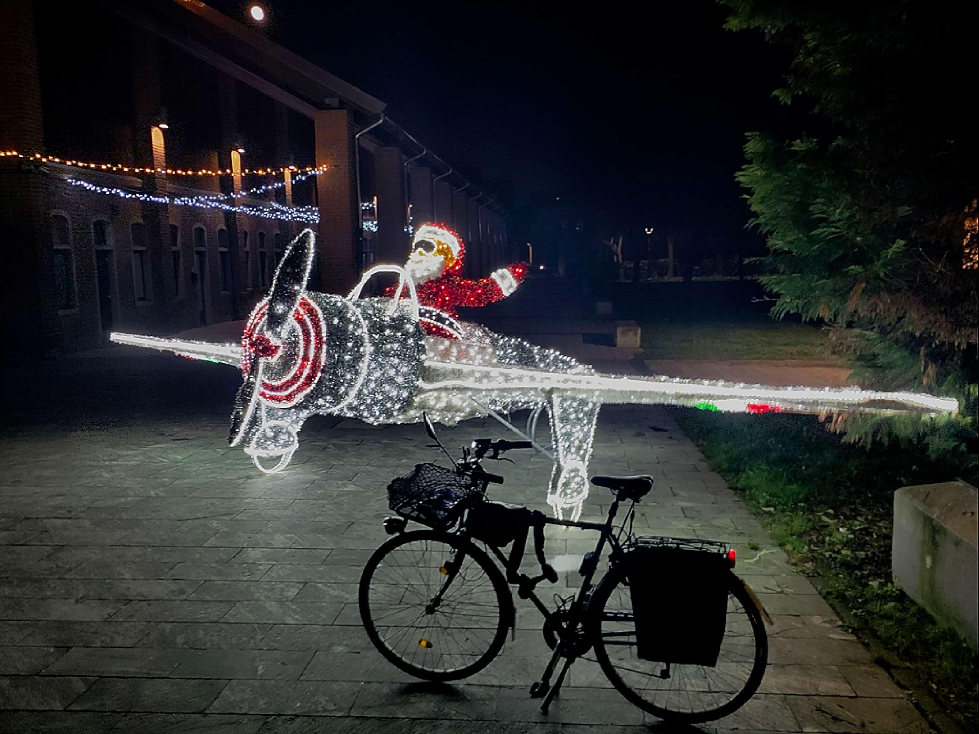 Foto di lato della mia bici blu. È notte. Dietro alla bici c'è un aereo fatto di luci (soprattutto bianche) di natale largo circa 8 metri, con rlics centrale. Il pilota dell'aereo è un babbo natale fatto di luci bianche e rosse. Dietro l'aereo si intravede una cascina