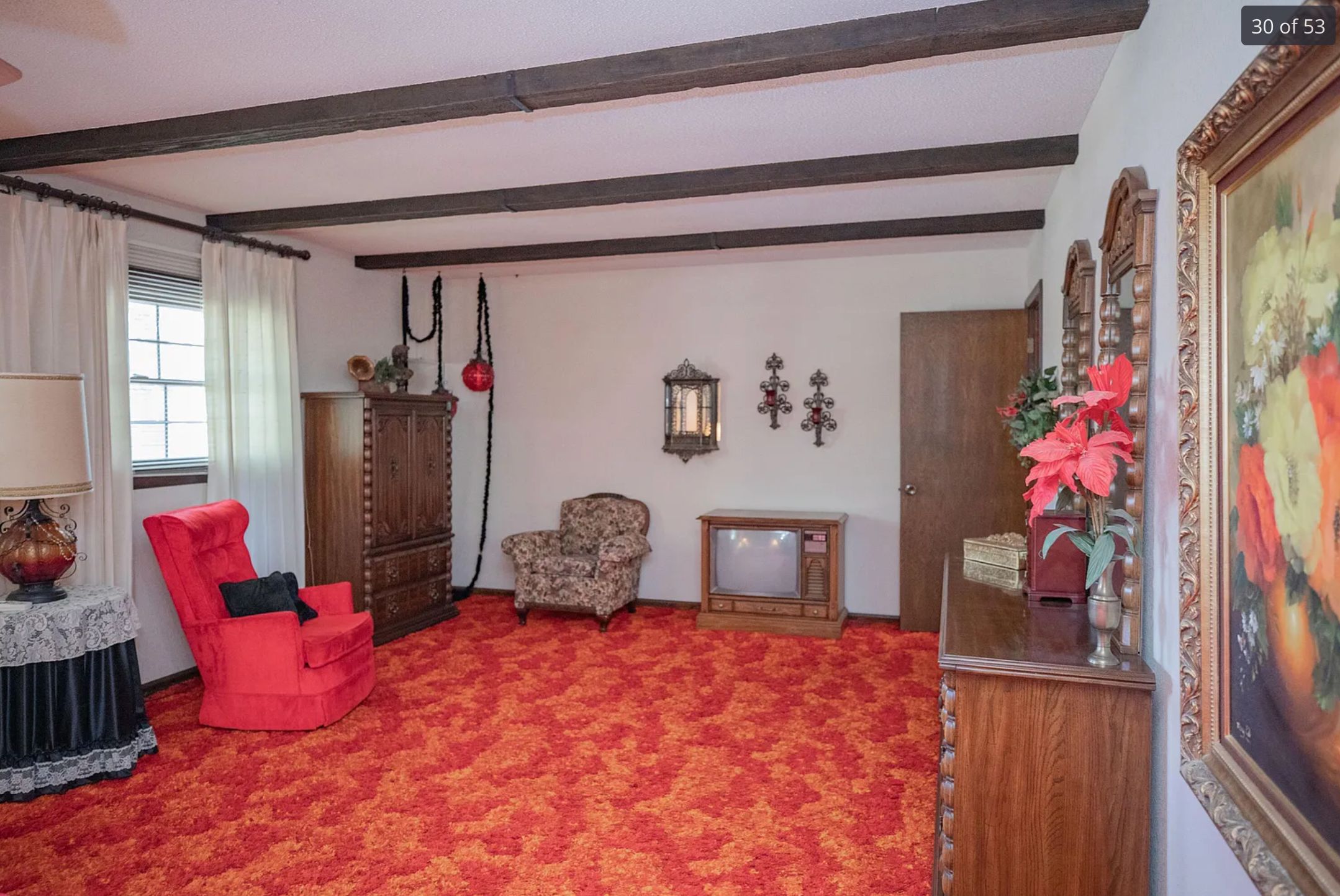 A very 1970s looking bedroom with an orange and yellow patterned carpet, and orange arm chair and a very old television set. There is also some artwork on the wall in gold frames.
