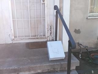 Photo of a white package under the black metal railing of some concrete steps in front of a house with white walls, a white metal screen door, and a white front door.