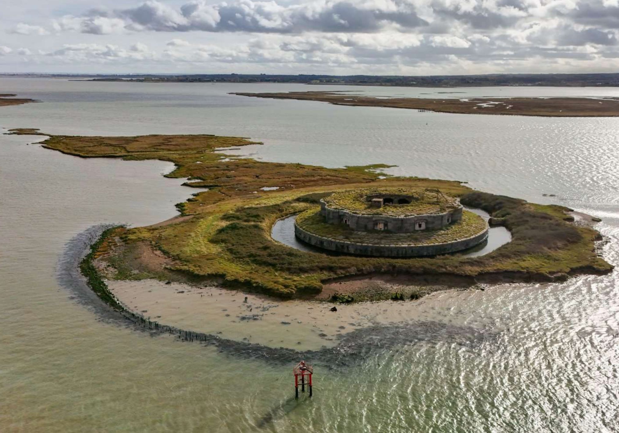 A photo of an old sea fort out on the water, covered by green growth and surrounded by patches of green land.