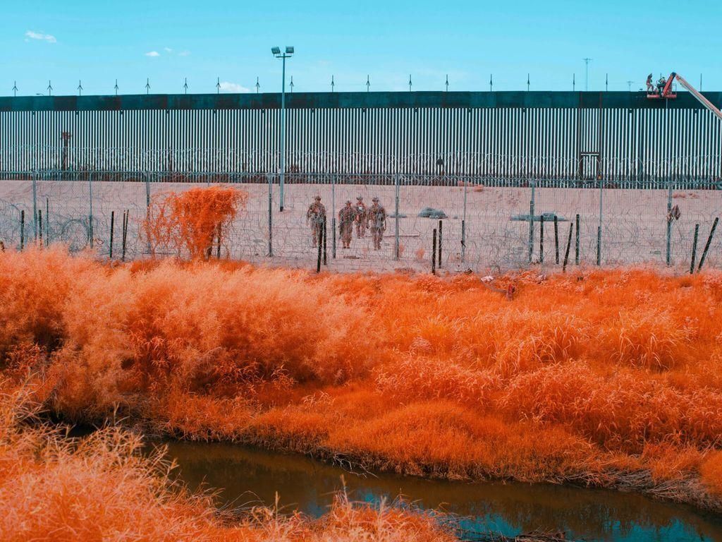 Seen in infrared light, foliage glows bright red while a cluster of troops beyond the border's barbed-wire wall are highlighted in brown. A tall border barrier stretches across the horizon behind them.