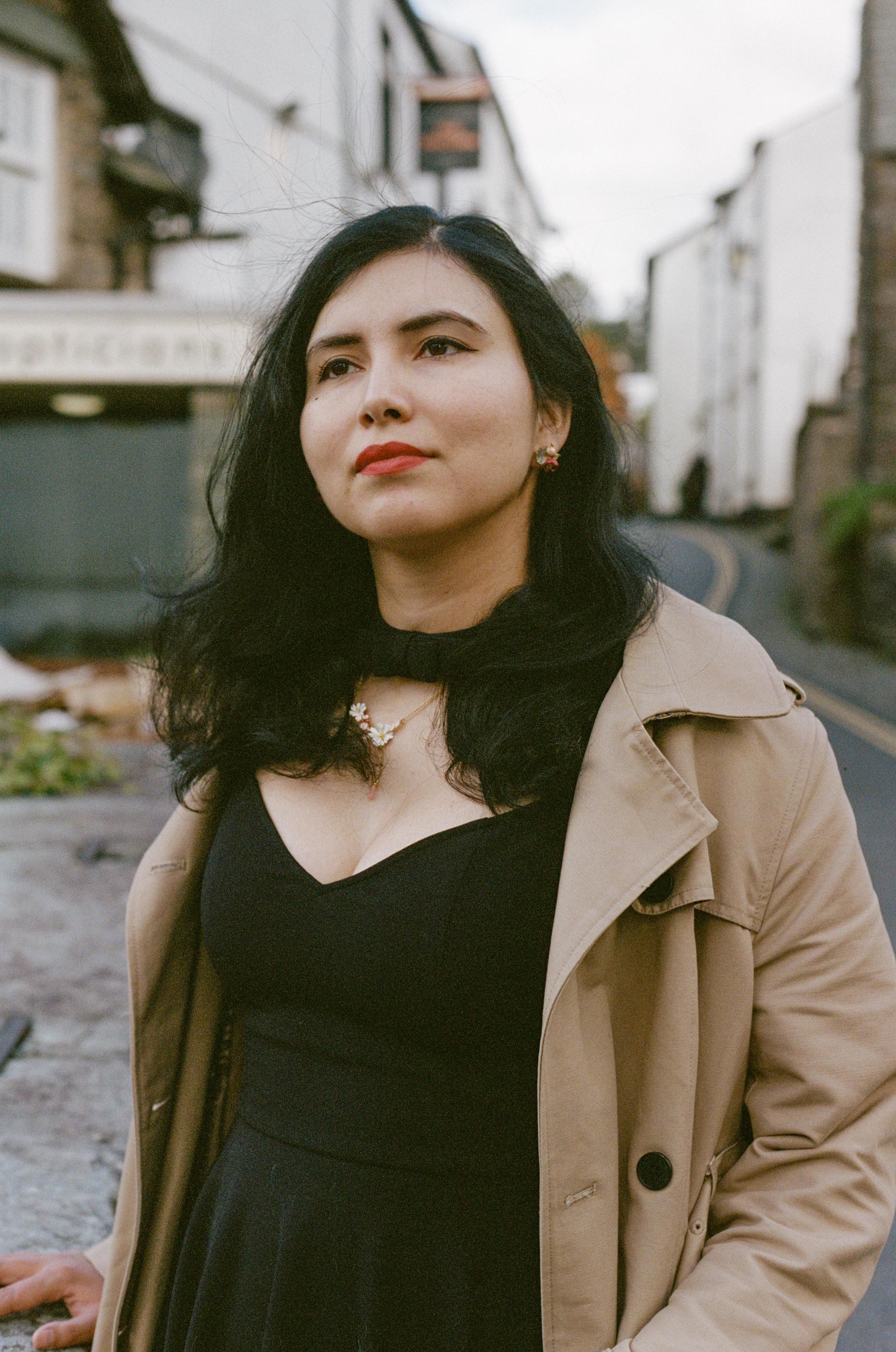 A beautiful woman with dark hair that has a slight blue sheen, wearing red lipstick, a floral necklace, a black dress with a low cut and a collar, and a beige trench coat on top.