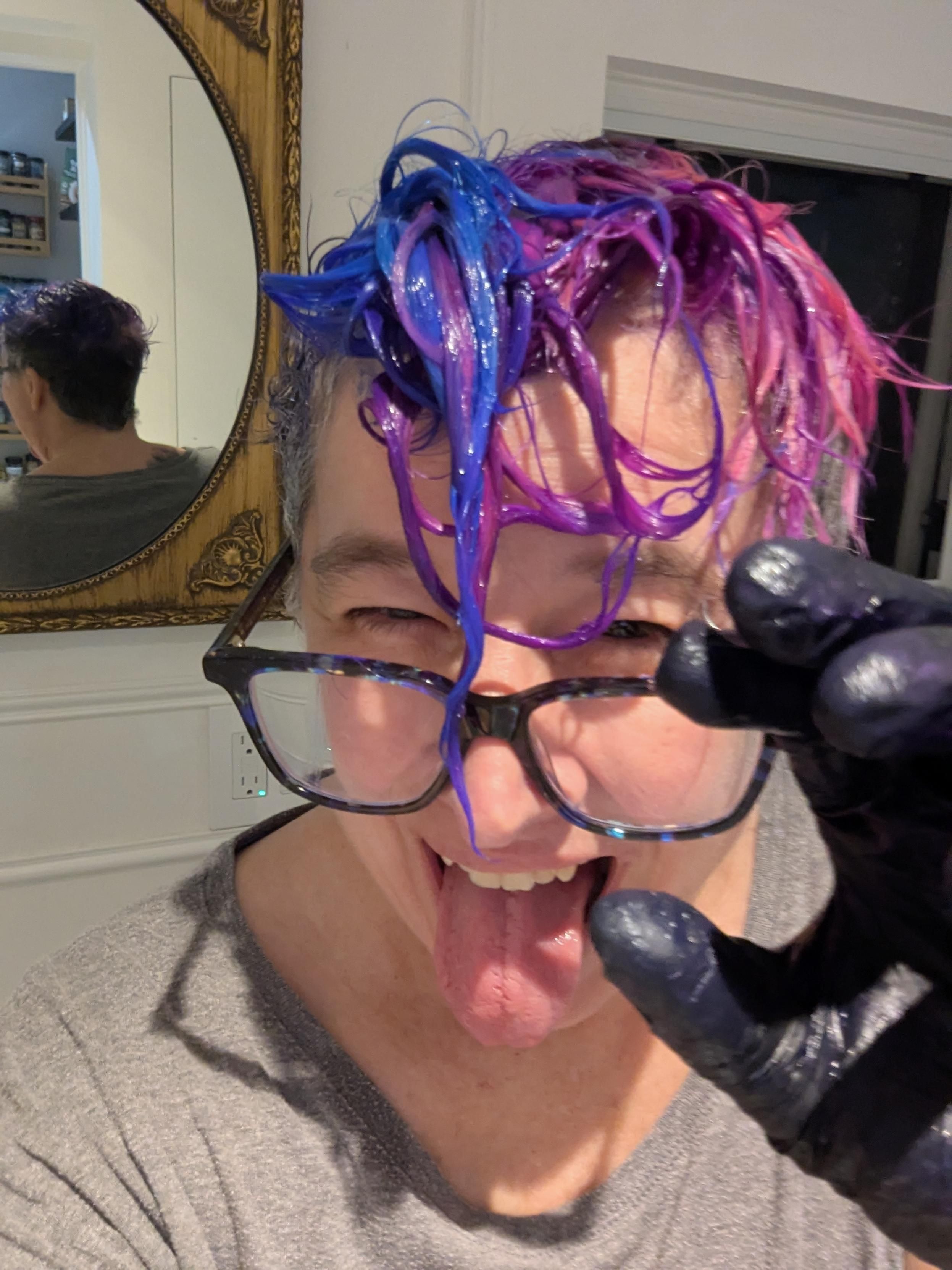 Blue purple and pink wet dye dripping off lizs hair into their face  one black gloved hand threatening the camera