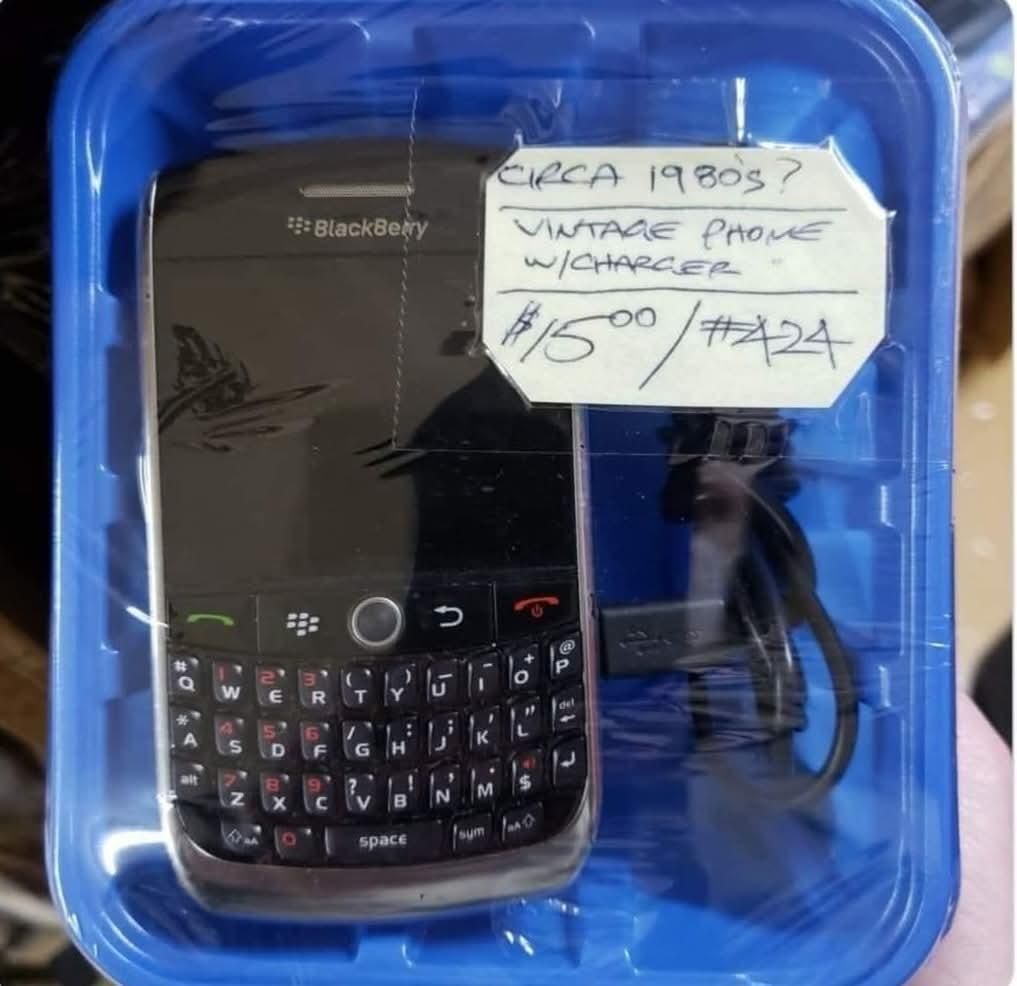 a blackberry smartphone in a blue box that feels like garage sale stuff
there is handwritten text on top that says "CIRCA 1980S? VINTAGE PHONE W/ CHARGER $15.00/#424"