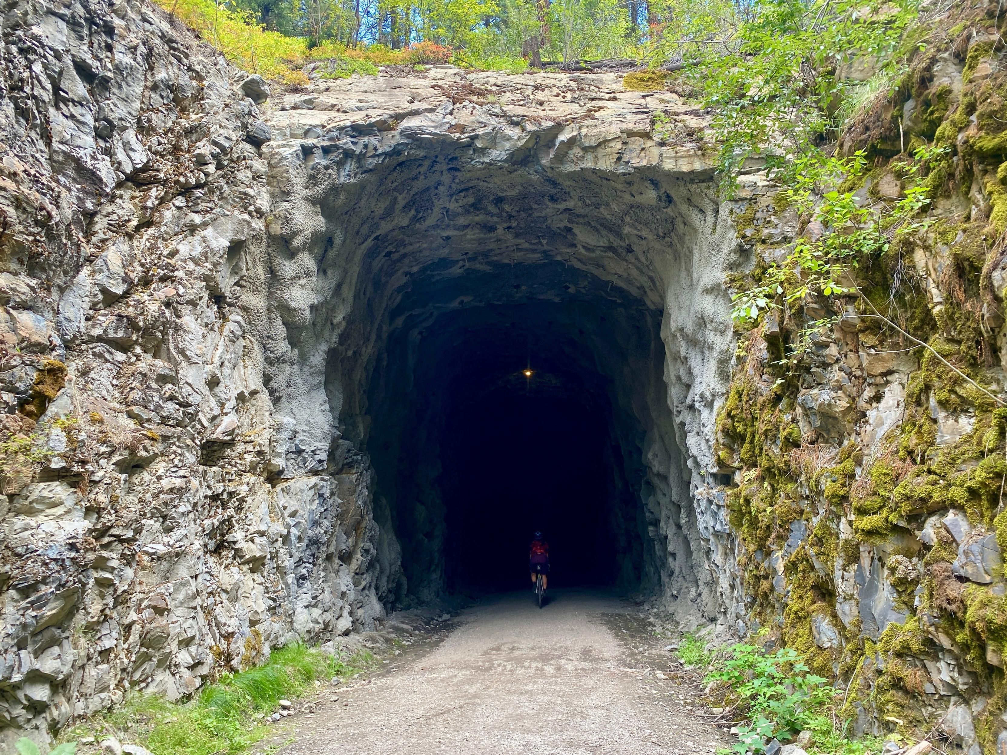 An above average sized make cyclist rides into a train tunnel that makes him look absolutely minuscule. He is barely visible, riding into the darkness. 