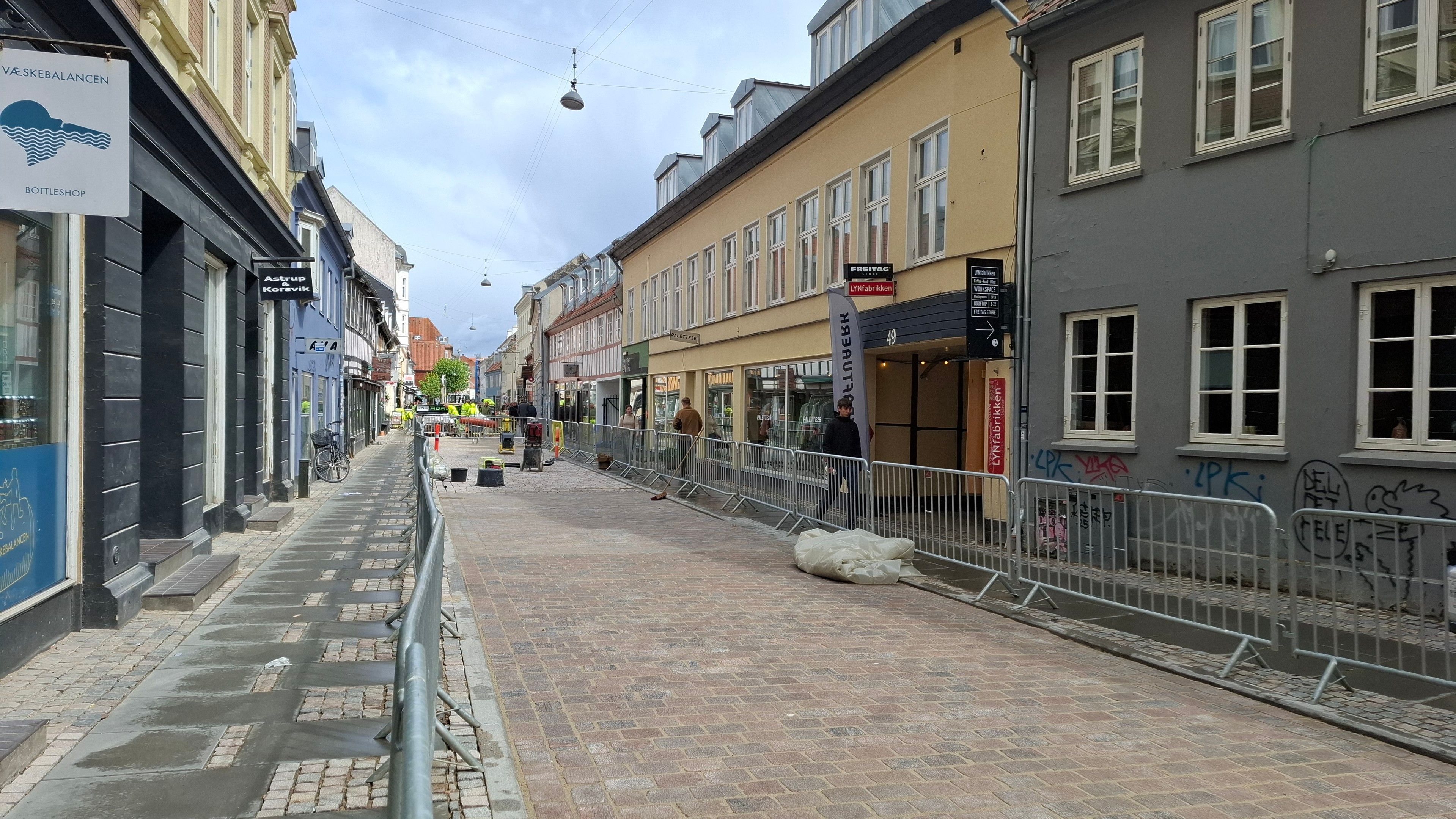 A finished section of the 'printed' brick surface - a long stretch of perfectly laid bricks. On both sides: small businesses and cafes and cyclists and pedestrians on the pavement alongside. Same general description from first photo now follows, so skip if already heard: Vestergade, mid-city Aarhus, Denmark. A lovely narrow old street that is, after a few years' trial, being turned permanently into a Quiet Street - cars may drive here, but only slowly, and bikes and pedestrians have priority. The council is planting trees, widening the pavement café and seating spaces, and laying a nice new chessboard-like brick surface. The surface is designed to slow down bikes too.  It is the first place in Denmark that is being paved with a Dan Jord RoadPrinter - a Dutch brick paver that rolls out complete stretches of road from a machine, as if you are winding a continuous sheet of paper out from a huge roll of paper. Photo taken with permission - I have turned into one of those Auld Lads who stand there, hands behind my back, watching them at work, fascinated by the process, interrupting them with questions including 'may I take some photos?