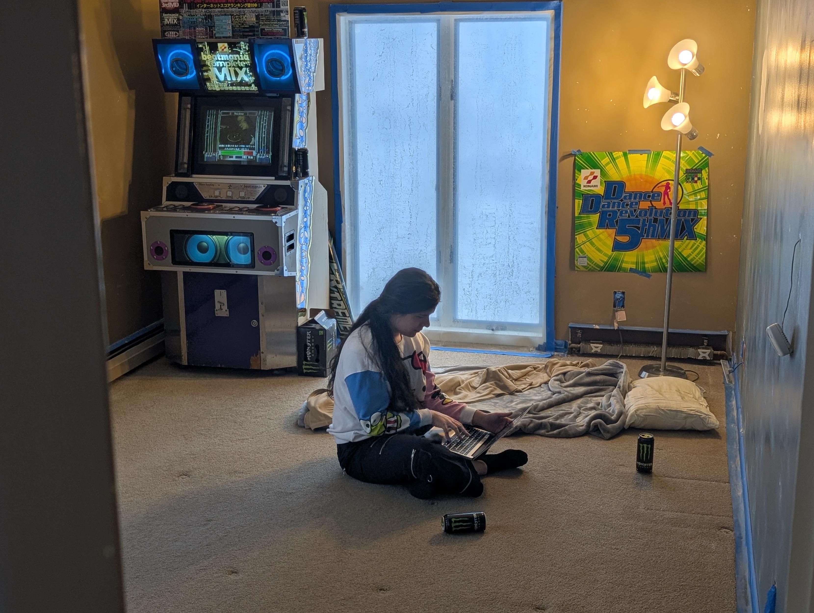 kira sitting on the floor of a mostly barren room using a laptop. there's a beatmania mini arcade cabinet running complete mix, a pillow and blankets on the floor, numerous monster cans, a DDR 5th marquee haphazardly blue-taped to the wall, and a bootleg Extreme marquee rolled up leaning on the cab. the walls are covered in that stuff you use to fill holes and there are numerous fixtures hanging out of one by their wires, the room looks under construction. the cab is in good shape and is very complete, with matching marquee and top pop, working lights, and its original crt. kira is wearing hot topic pants and a hello kitty sweater 