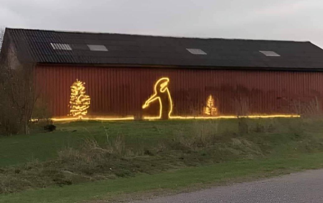 A rural red barn decorated with a glowing yellow light installation showing a human silhouette urinating between two illuminated Christmas trees, with a continuous line of lights running along the ground. The humorous outdoor light artwork contrasts festive holiday symbols with crude humor, set against a grassy roadside landscape at dusk. Artist unknown, location unknown.