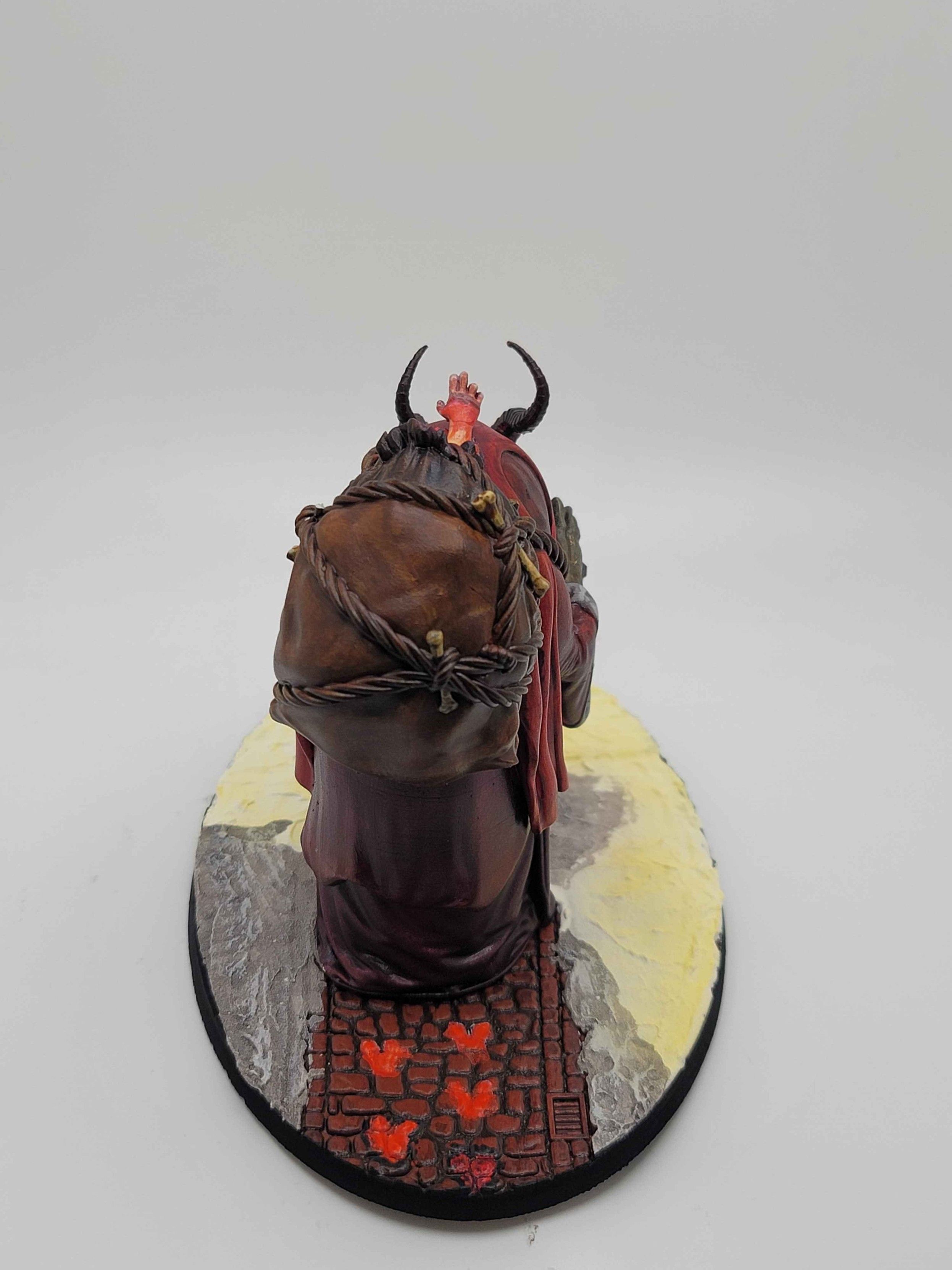 The rear of the same miniature, showing the harsh shadows cast by the lantern, and the child's arm lit from this angle by the fires of hell within the sack on Krampus's back.  The road behind Krampus is not snowy, all the snow having been melted by his hellish heat, as he left glowing hoofprints on the cobblestone road, revealing that his feet are the cloven hooves of a demon.