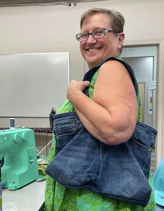 A woman holding a denim tote bag made from a pair of jeans. A teal green sewing machine sits in the background.