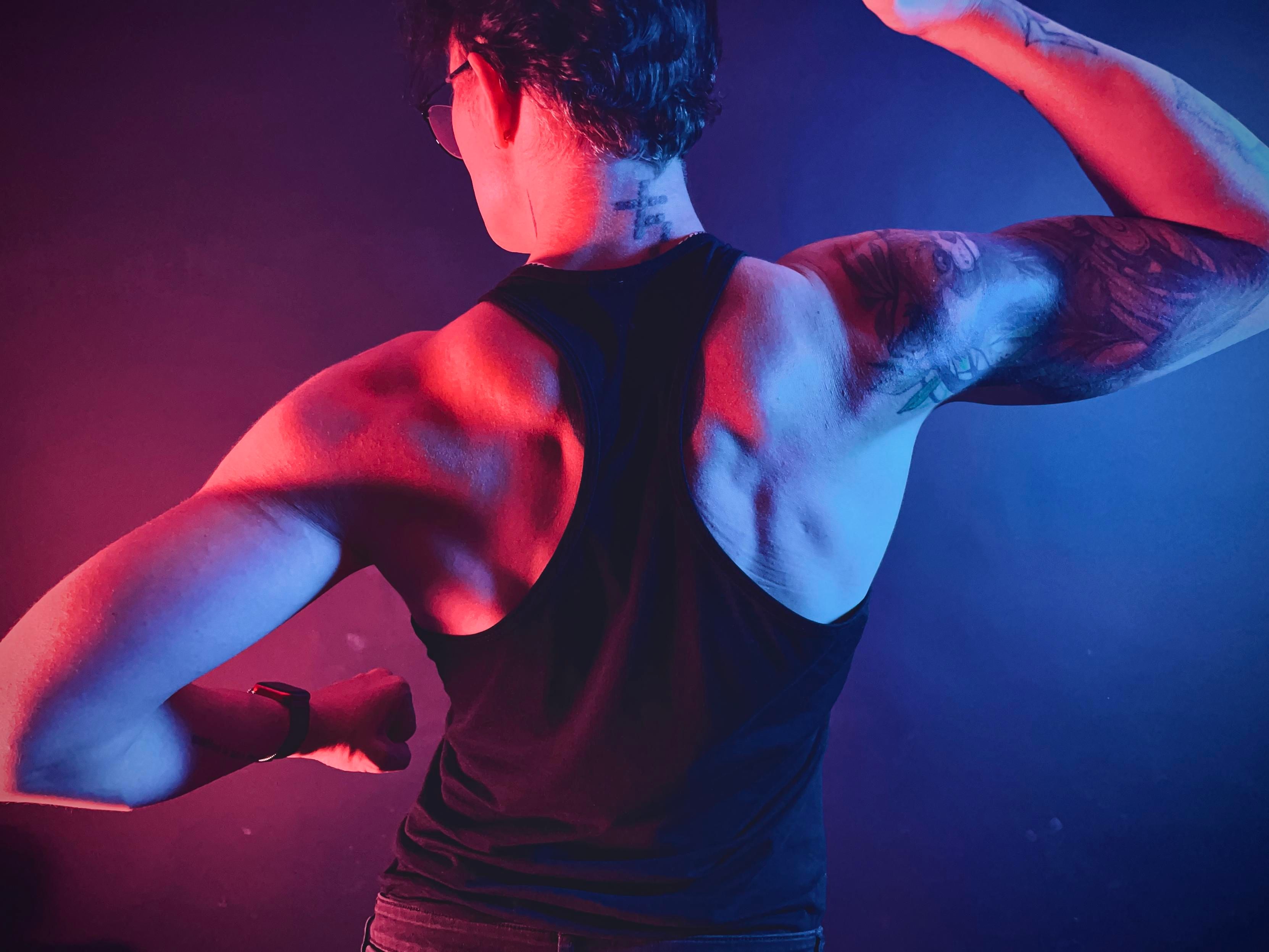 Photo of my back flexing from behind, wearing a black racer tank top, lit blue from the right and orange from the left, emphasizing the back muscle definition 