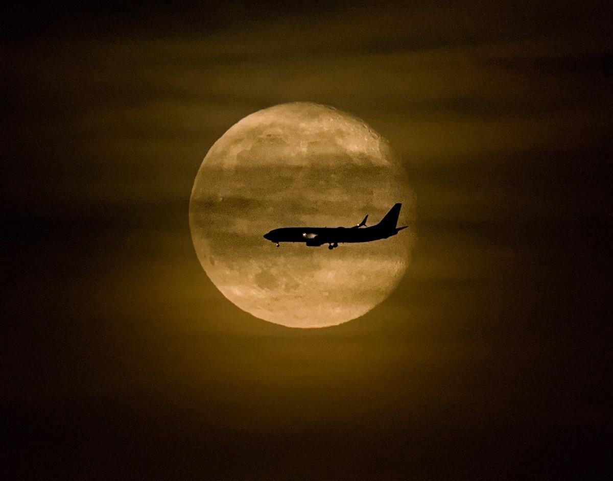 A color landscape photo of a jetliner passing in front of a full moon. The moon is a light-yellow and a bit shrouded in hazy thin clouds. The thin clouds extend out past the moon's edge like a painter's bush strokes. 
