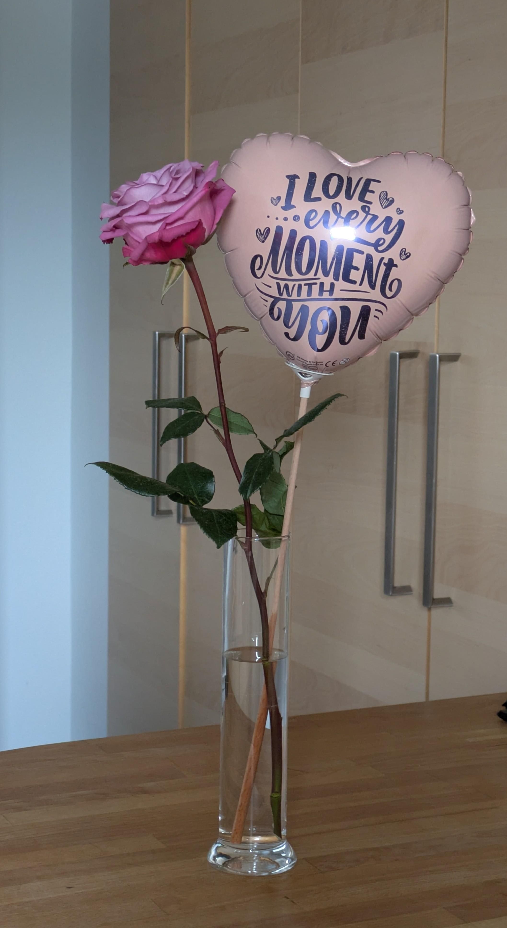 a pink rose and a nearly perfect color matching balloon in heart shape with the text "I love every moment with you"
both in a vase