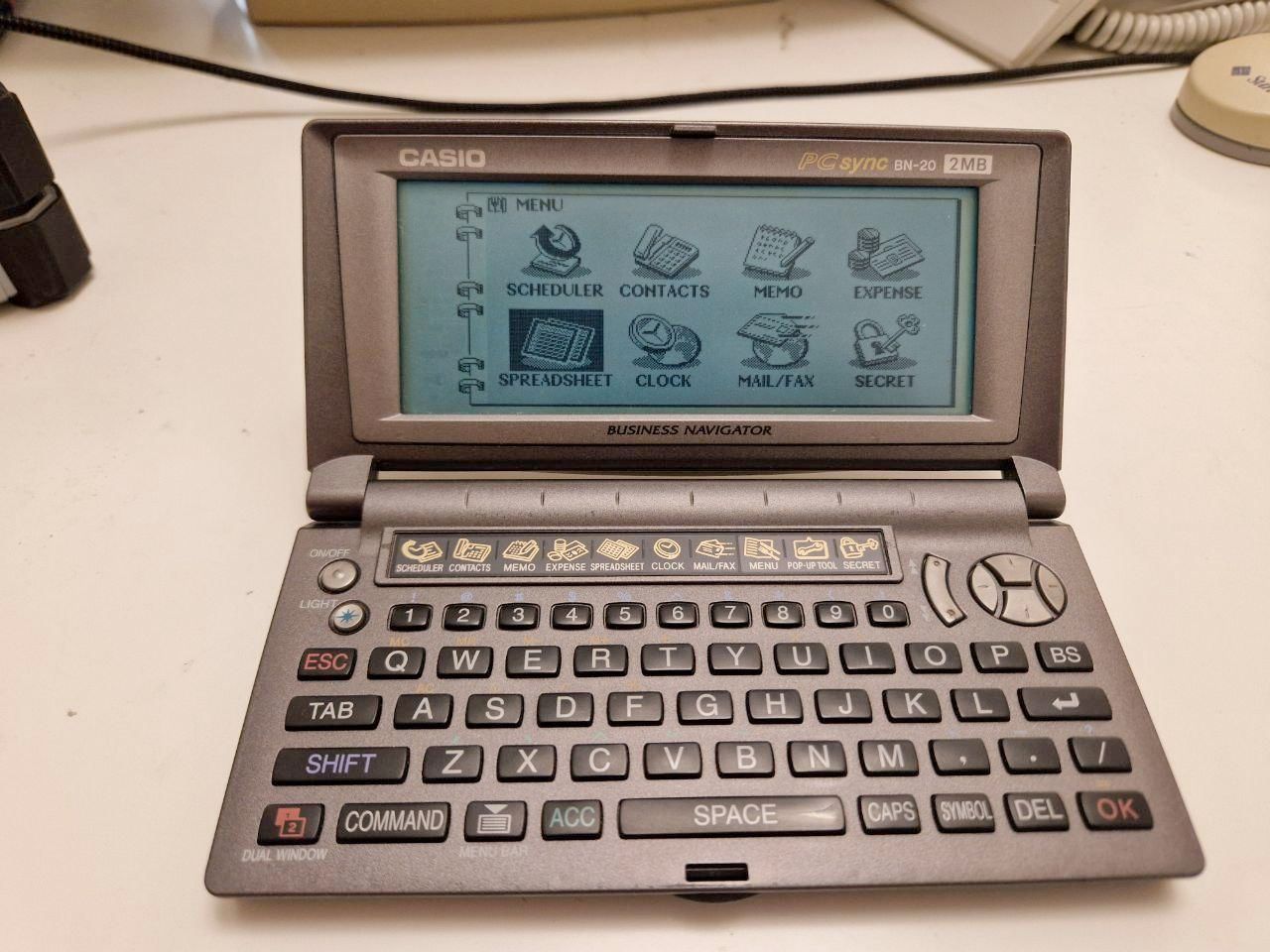 A photo of a tiny pocket organiser with a QWERTY keyboard, showing the main menu (calendar, scheduler, spreadsheet, etc) on its monochrome screen