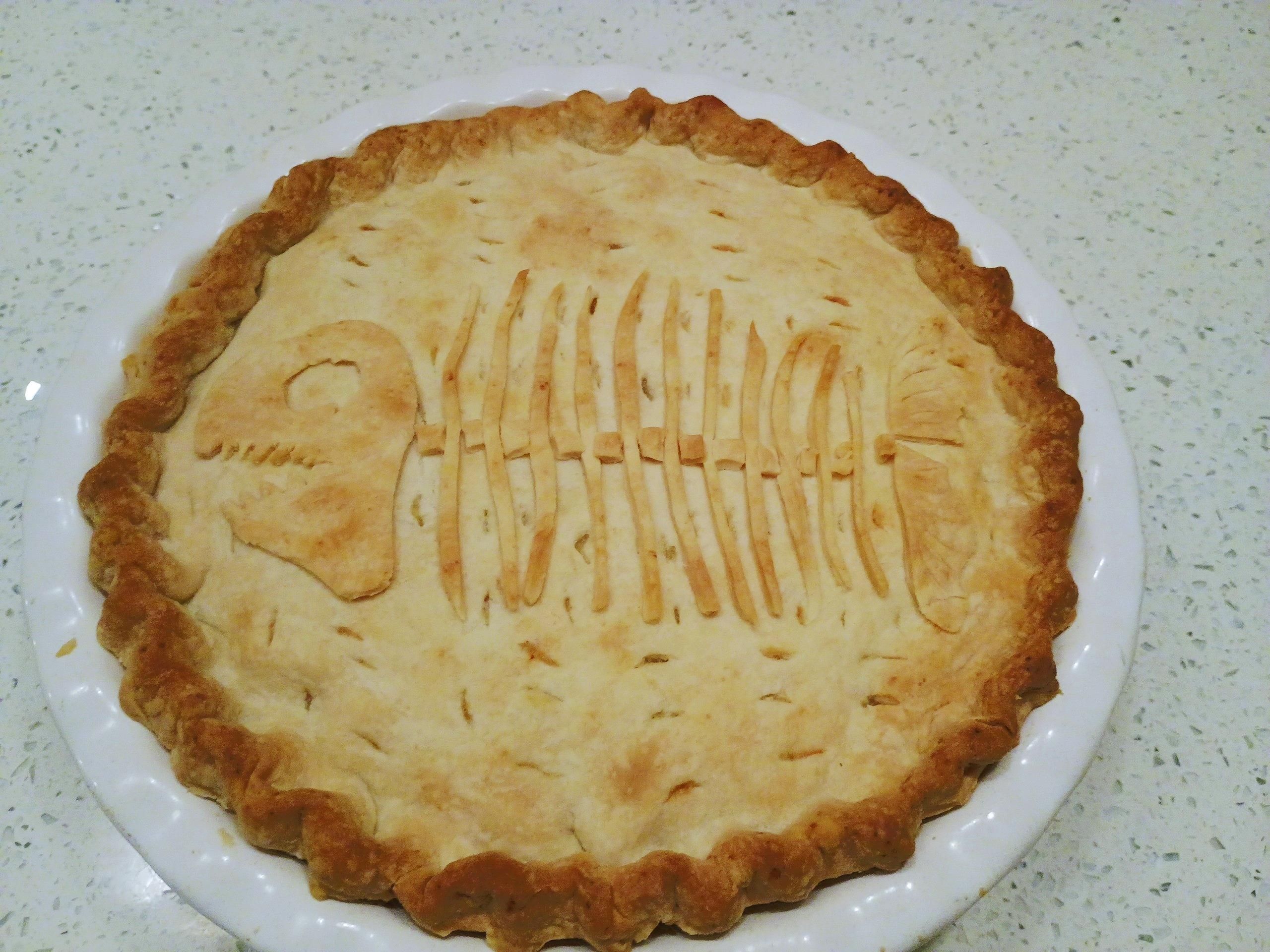 Golden brown pie in a white deep pie plate features a decorative fish skeleton on top and a crinkly crust.