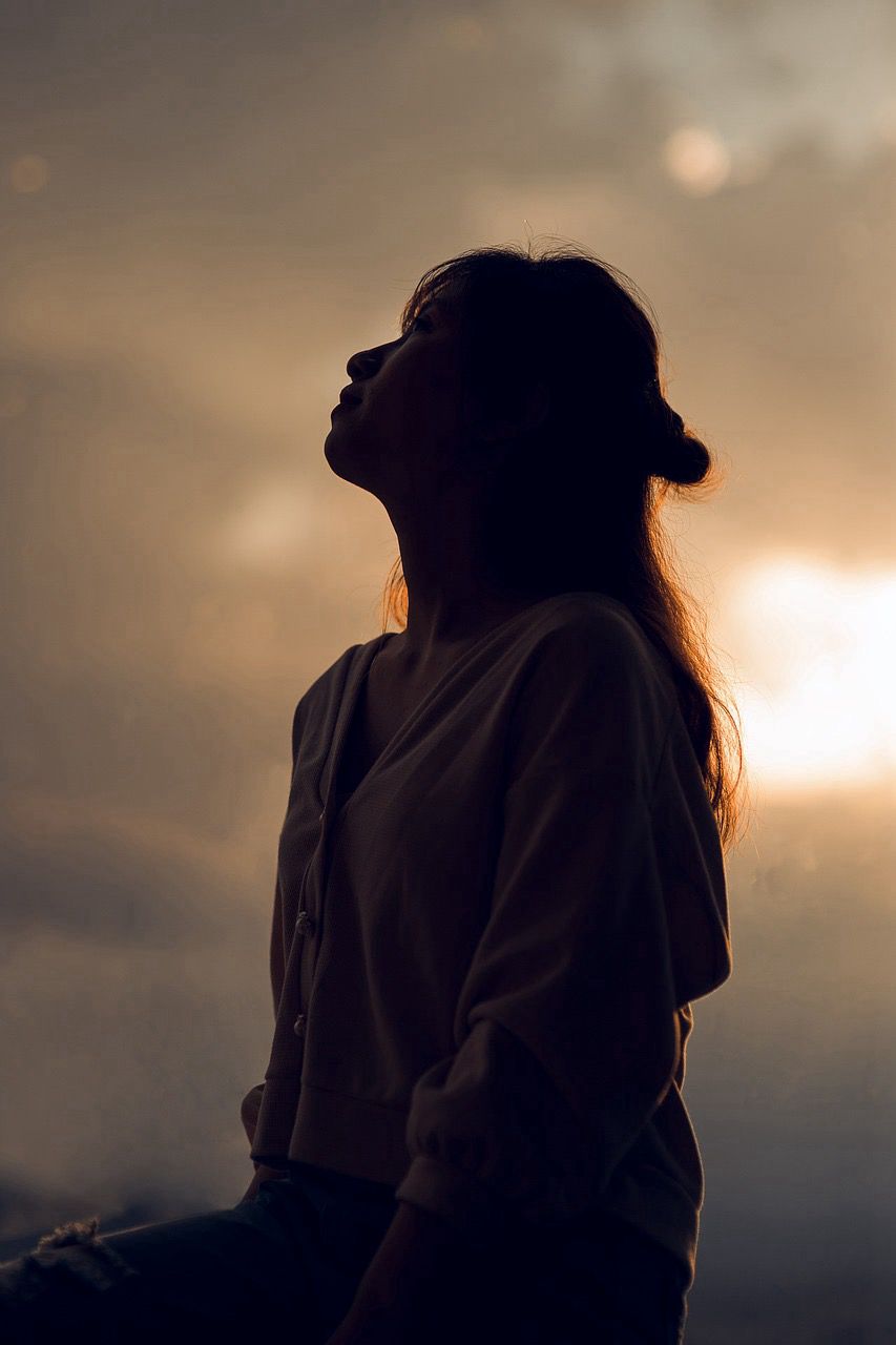 A woman looking to the sky silhouetted against a sepia background, a bright light behind her.