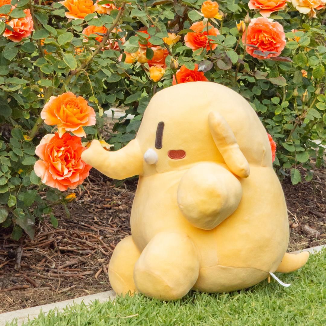 Photo of the Creature Mastodon stuffed toy sitting at the base of a rose bush and smelling some flowers during a bright afternoon. The Creature is on the concrete edging separating a mulched rose bed and a lush green lawn, its body perpendicular to the camera and facing image-left. Its trunk is pointed directly at two low-hanging Rosa “Piñata” blossoms.