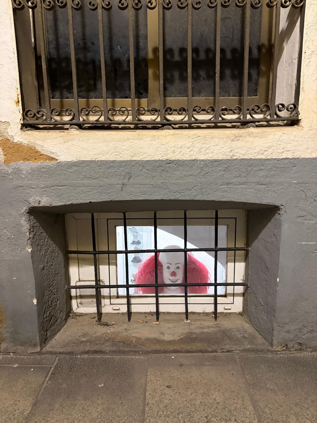 Barred basement window with a mural depicting a fake clown with red hair and white face behind it. The wall is weathered, creating a slightly eerie mood.