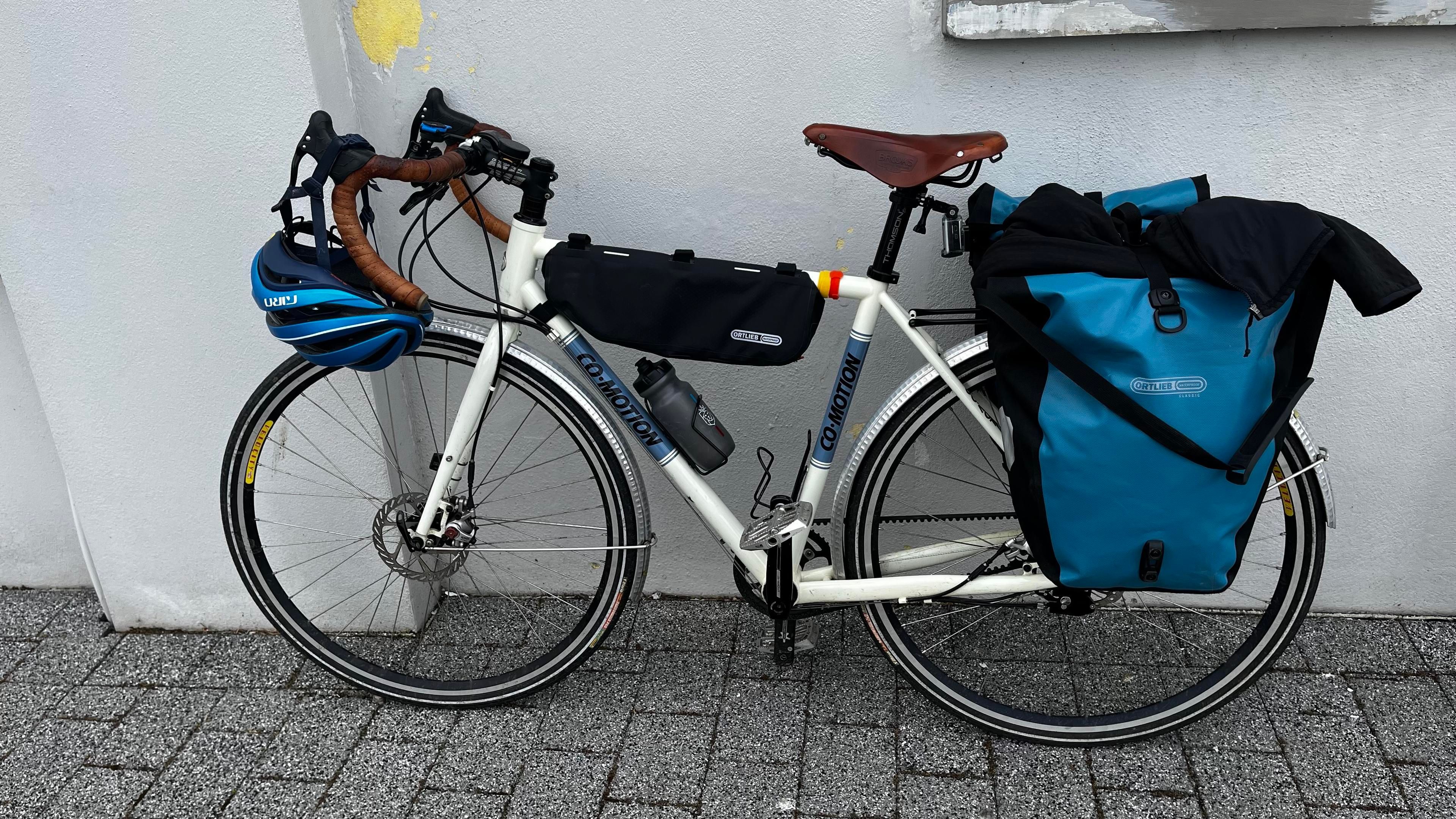 A white bicycle with a brown leather saddle and handlebars is parked against a gray wall. It features a black frame bag, a water bottle, and a blue helmet on the handlebars. Two blue pannier bags are attached to the rear of the bike.