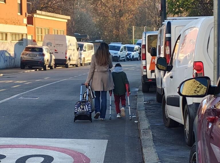 Foto di una strada. I marciapiedi sono occupati da veicoli in sosta, costringendo una mamma e un bambino camminano in carreggiata. La mamma porta lo zaino trascinandolo sulle ruote, il bimbo cammina con le stampelle. Si vede parzialmente sull'asfalto il pittogramma di limite 30km/h.

Foto dall'articolo di urbanfile