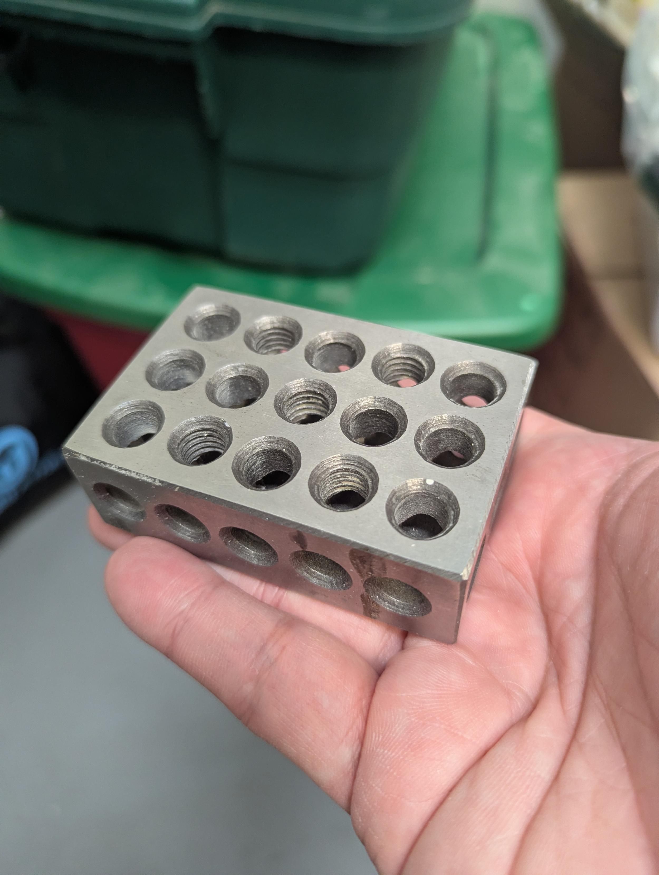 A metal box shaped object with many holes in it. It sits in the palm of a hand. It's original use is for machine tool setup but this one is beat up and no longer useful for that. 
