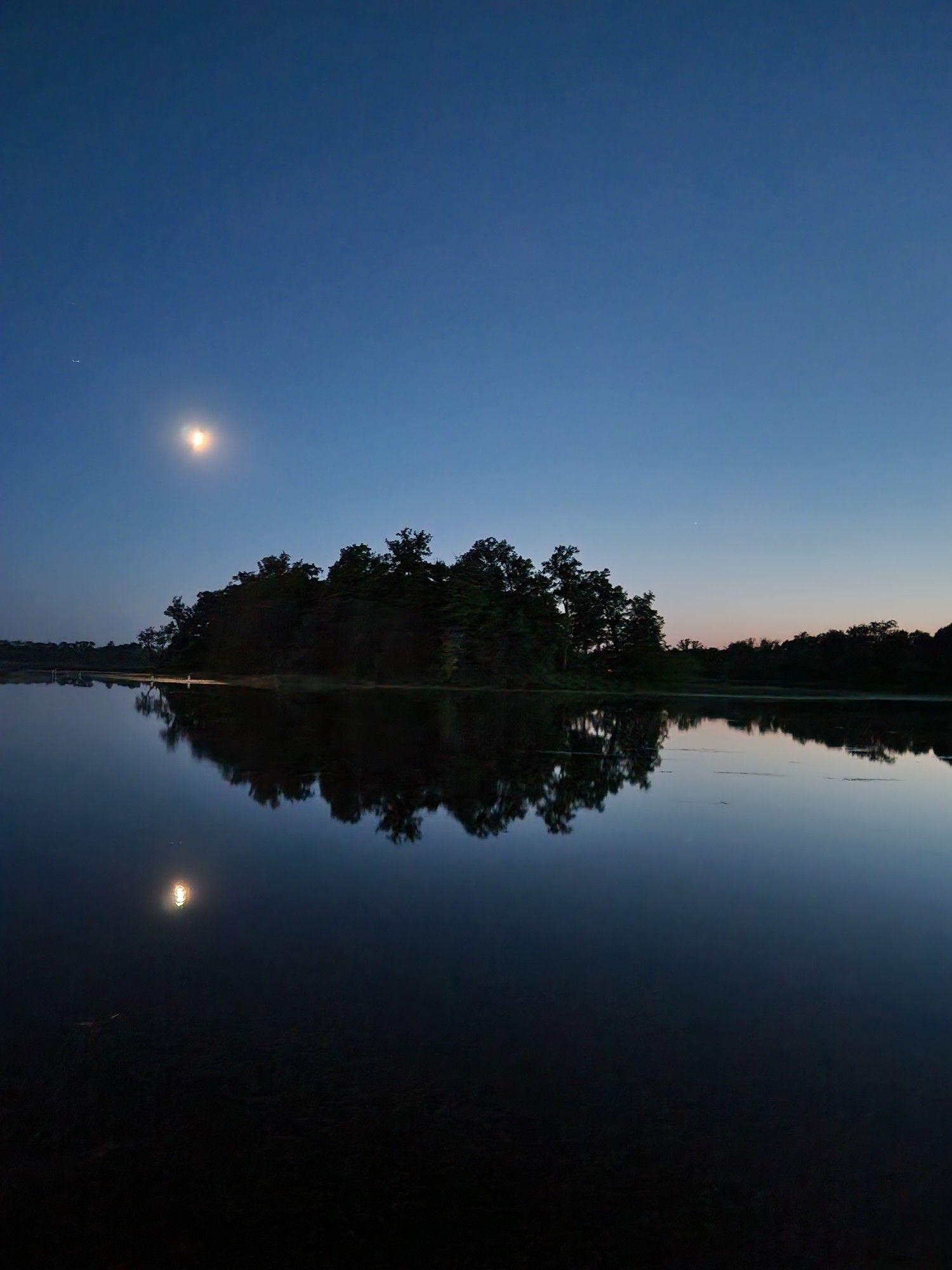 The moon shines over a calm lake at dusk, with an island in the middle of a lake.