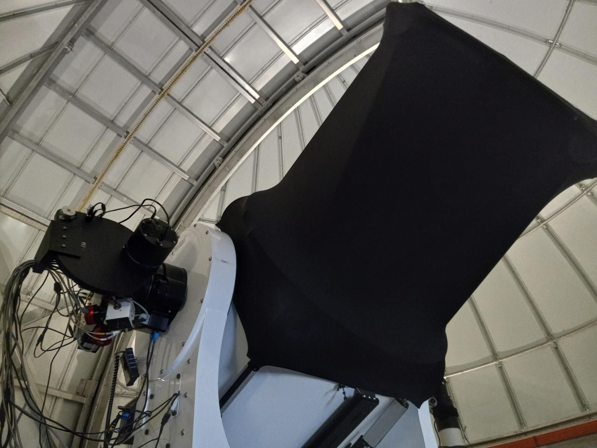 Looking up at a very shiny new large telescope white telescope with a black dust shield and a lot of wires and camera equipment on one side, inside a white dome.