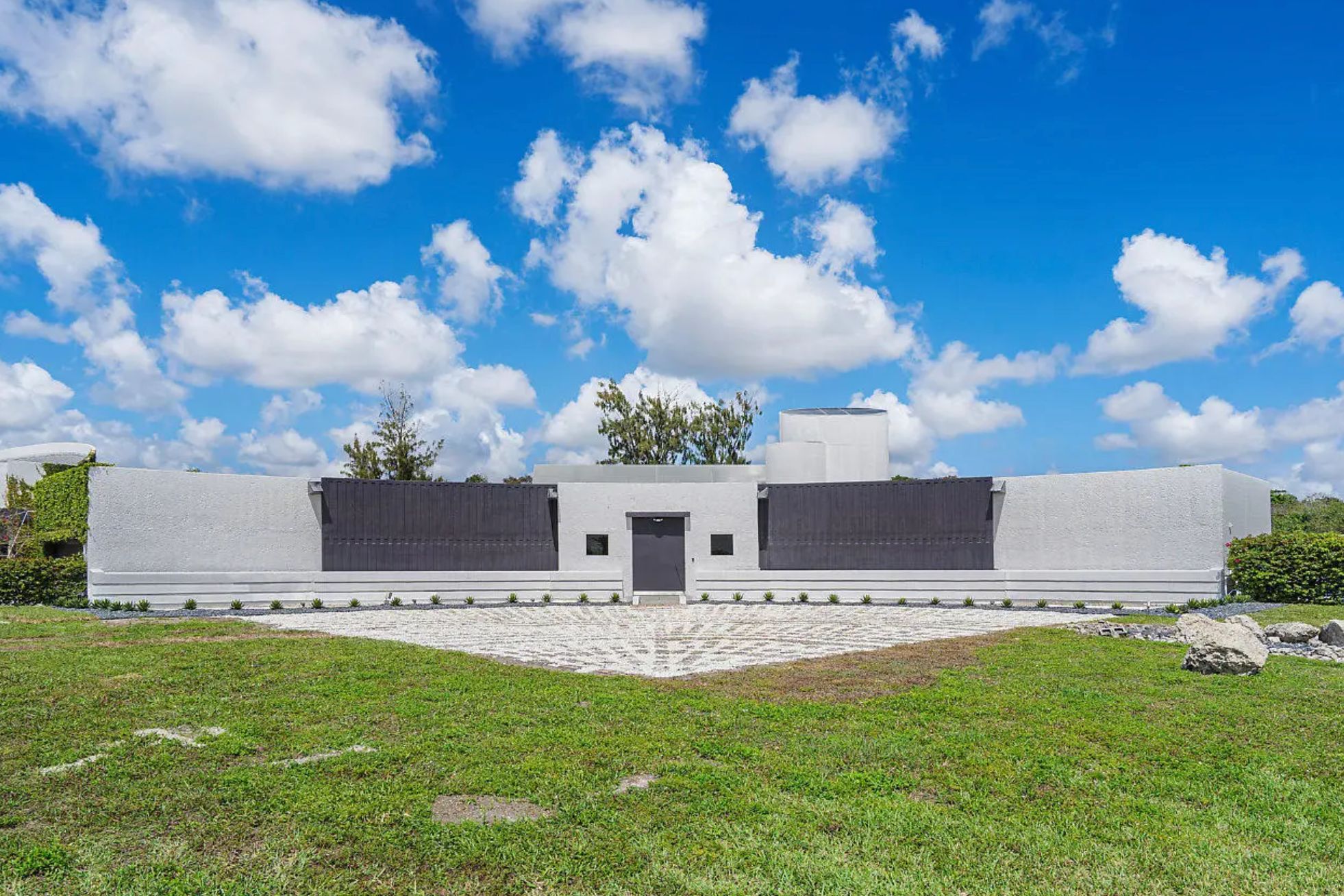 An one story curved structure that is flat on top and is concrete except for a front door flanked with small square windows, and dark grey panels. There is a triangle area in front where one could presumably park a car, but no driveway coming up to it. 