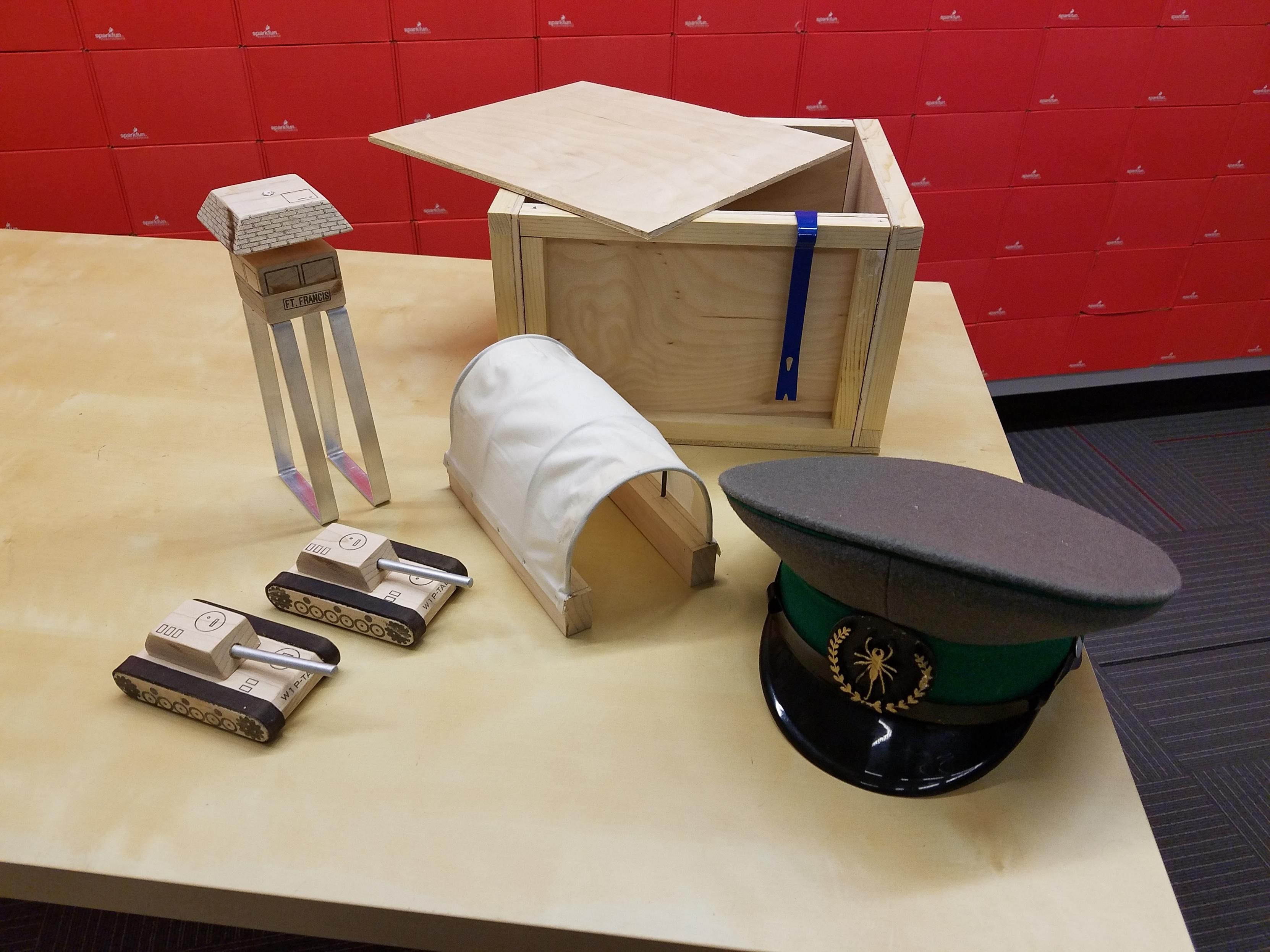 The contents of the crate spread out on a table. Two plywood tanks with leather strap treads, a watchtower on an aluminum base, a canvas tunnel, and a General's hat with a 3D printed vinegaroon insignia