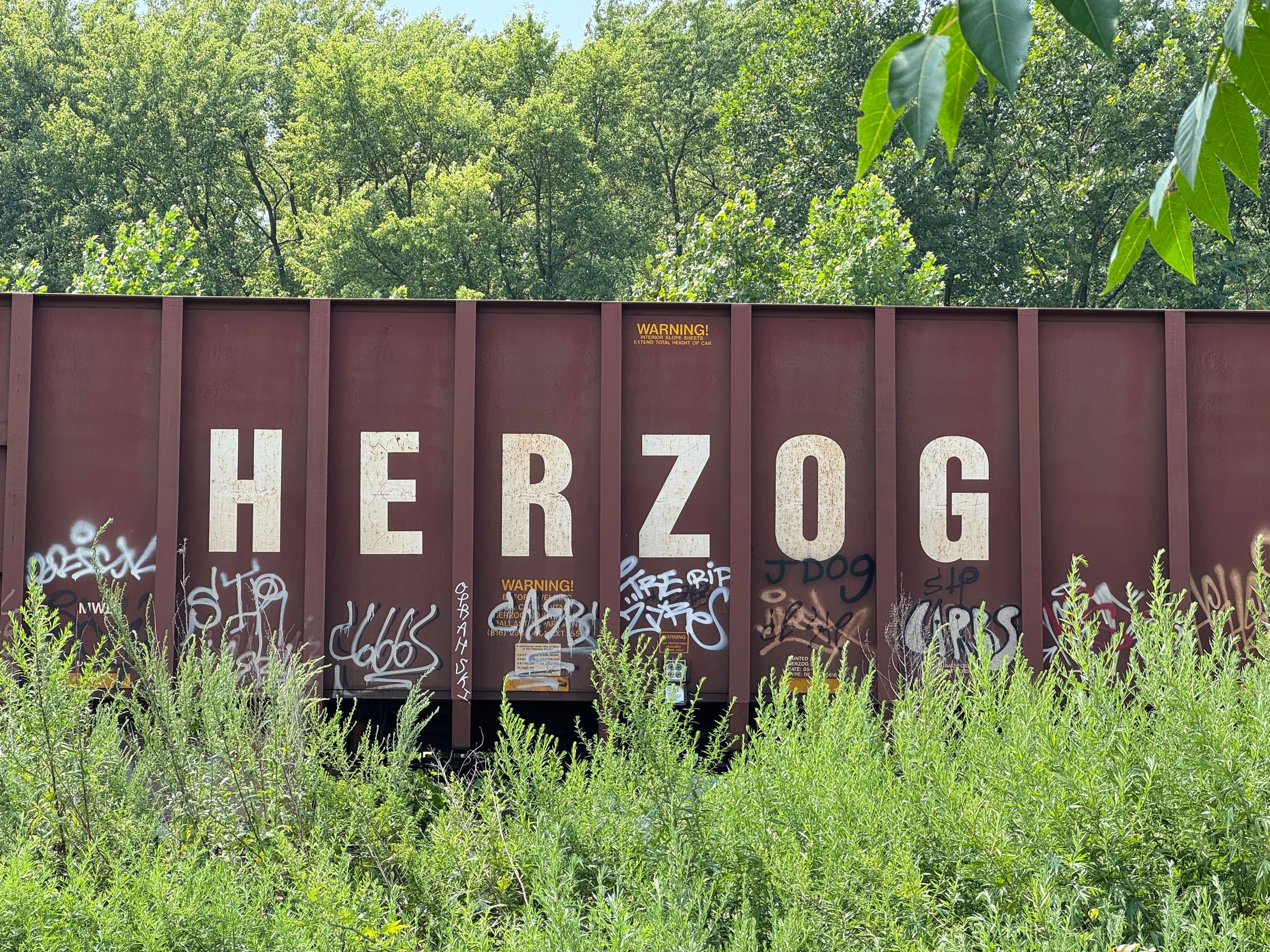 A train car with ‘HERZOG’ in large letters on the side. In very small yellow letter me above it says ‘Warning!’ There is graffiti at the bottom of the train.