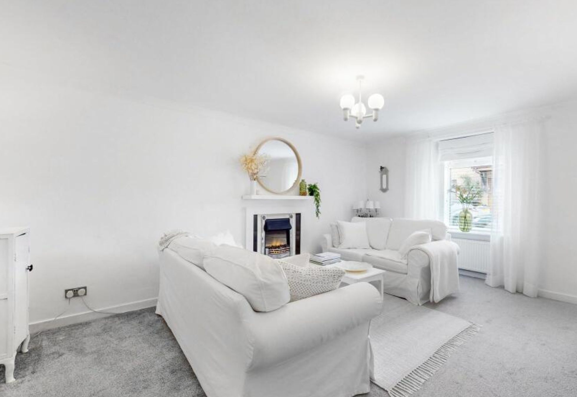 A living room with light grey carpet and bright white walls, ceiling, couch, curtains, throw blanket on the couch, and rug. There is a fireplace that is also white with a black accent and a gold mirror on it. 