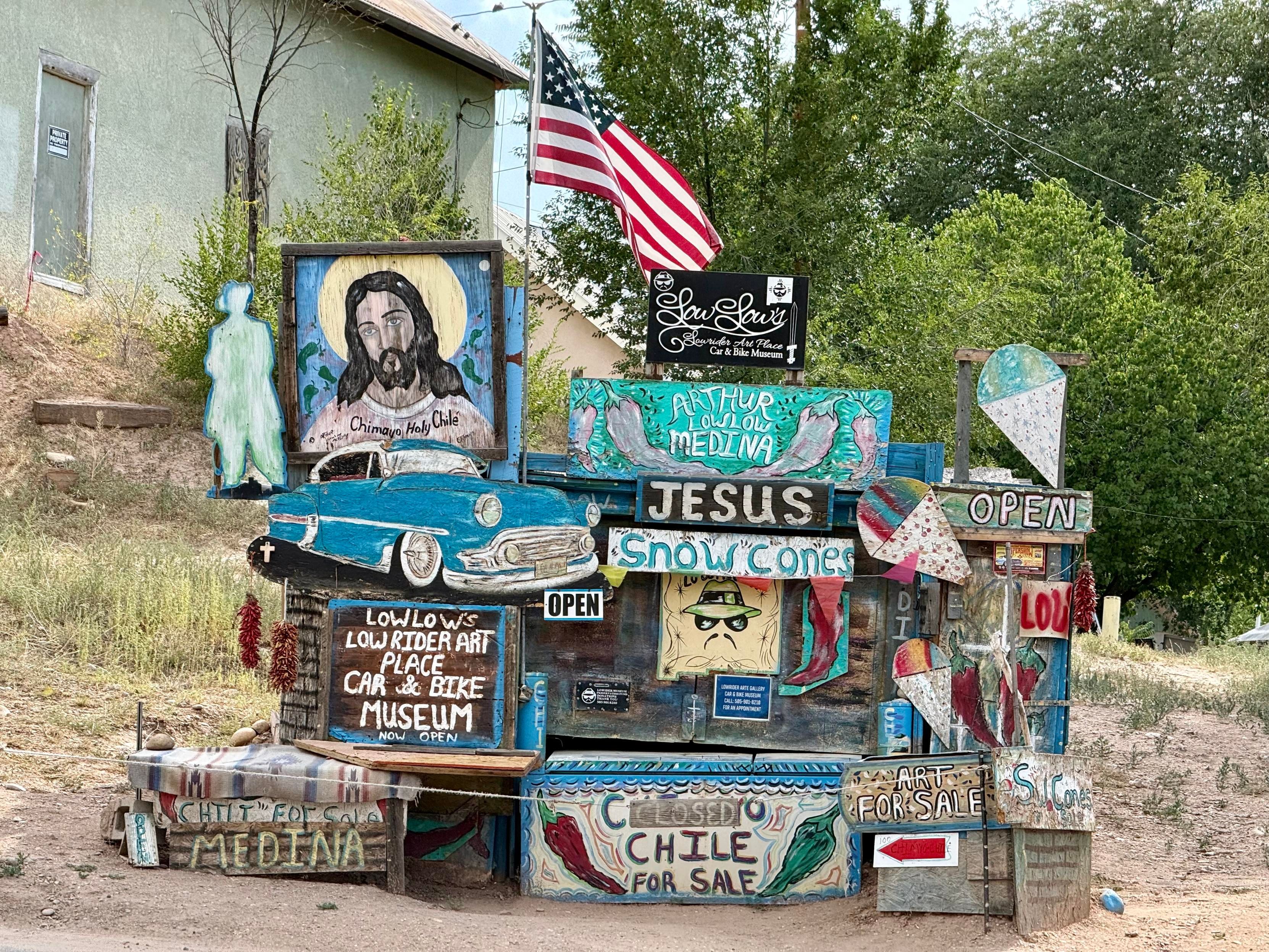 The image depicts a colorful, eclectic roadside attraction with various signs and decorations. The structure is made of wood and features a mix of painted signs, images, and objects. At the top, an American flag is prominently displayed. A large sign with the text "JESUS Snow Cones" is visible, along with a sign that reads "Arthur Lou Low Out Medina." A sign indicating "LOWLOWS LOWRIDER ART PLACE CAR & BIKE MUSEUM NOW OPEN" is also present. The structure includes a painted image of a blue car, a religious painting of Jesus with the text "Chimayo Holy Chile," and a sign that says "Chile for Sale." There are also signs that read "OPEN" and "Art for Sale." The background shows a building and trees, suggesting a rural setting. The overall scene is vibrant and filled with a variety of artistic expressions.

Provided by @altbot@fuzzies.wtf, generated privately and locally using Ovis2-8B

🌱 Energy used: 0.235 Wh