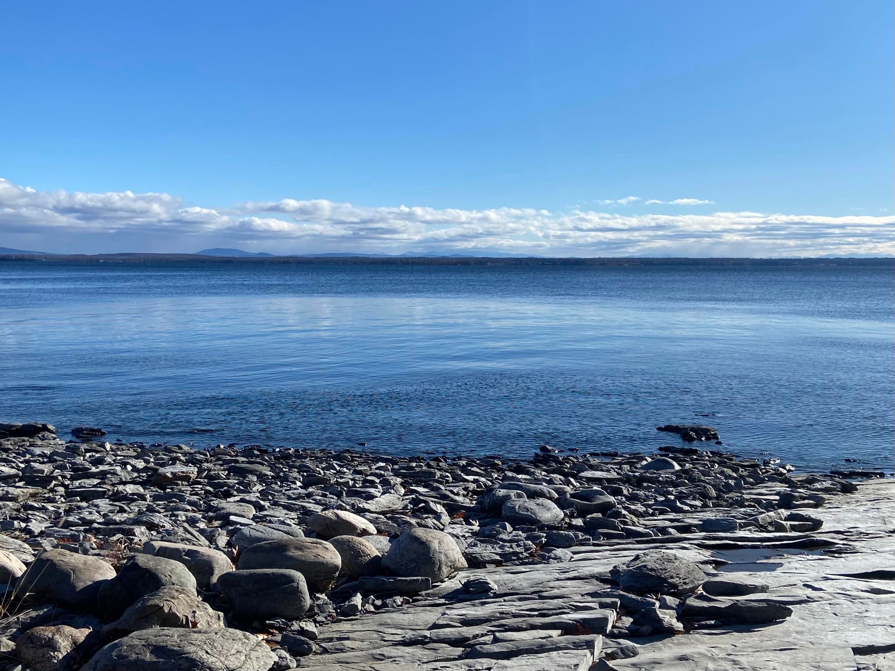 A gray rocky shore in the bottom third of the photo, deep blue lake in the middle third, and blue sky with a few clouds at the horizon in the top third. We're looking East across lake Champlain, standing in New York, looking at Vermont on the far shore. 