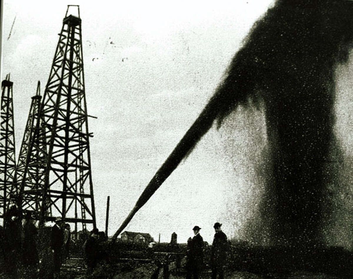 The Spindletop oil field gushing with a cloud of black oil, silhouetted against the sky, along with oil derricks and onlookers.