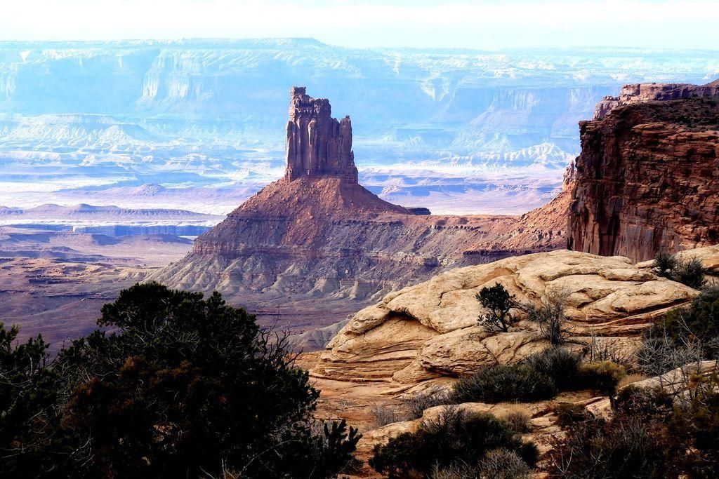 A tall rock formation rises from a layered desert landscape in Canyonlands National Park, with cliffs and mesas in the background. The foreground features rocky terrain and sparse, scrubby vegetation. The scene appears arid and rugged.