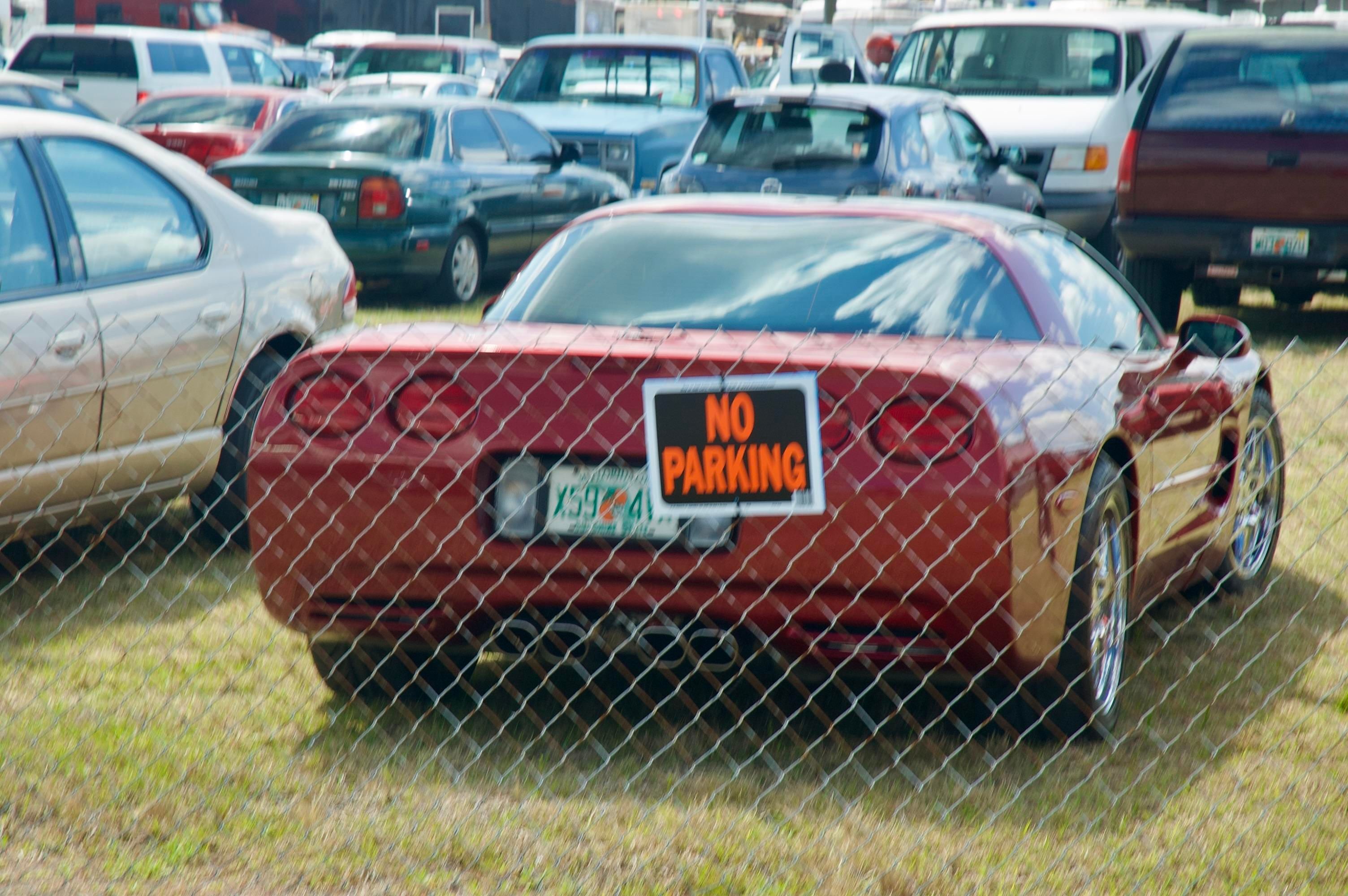 A wire fence with a NO PARKING sign. Beyond it, many cars are parked.