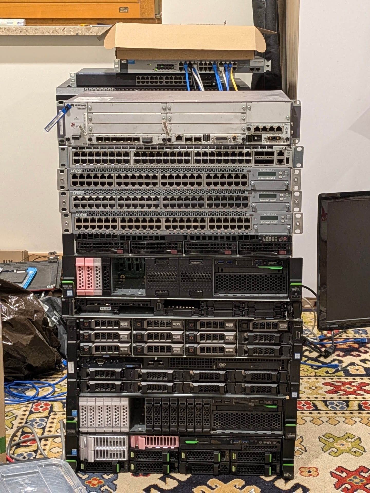 a stack of server hardware (top to bottom):
- Huawei BBU3900
- Arista DCS-7050TX
- 3x Juniper EX4300-48P
- Supermicro x11ddw-l
- Fujitsu RX2540 M5
- Dell R640
- Dell R730xd
- Dell R730
- Fujitsu RX2540 M1
- Fujitsu RX2540 M4

also a Procurve 1800-24G and an EX2200-24P in the background just above the main stack