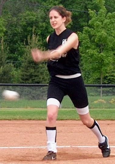 A picture of my daughter when she was 17 throwing a pitch in a softball game. She is wearing a black jersey and shorts, with white socks with black trim. She is in mid stride of her pitch, left-handed, and has just released the ball, which can be seen as a blur on the left side of the photo.