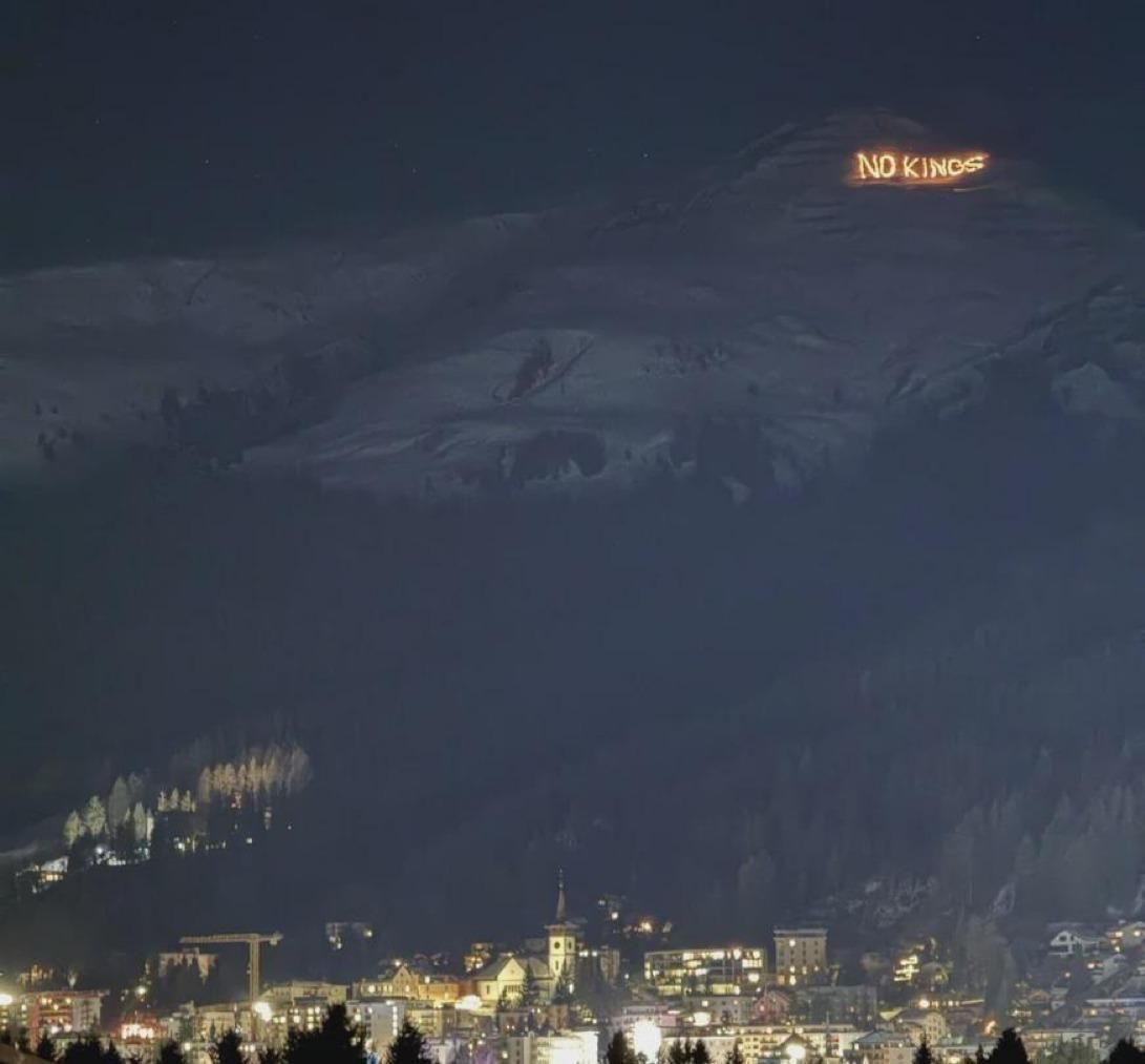 Nighttime view of Davos, Switzerland during the World Economic Forum, with the town’s lights below and a snowy mountainside above, where the words “NO KINGS” glow in bright letters on the ridge against the dark sky.