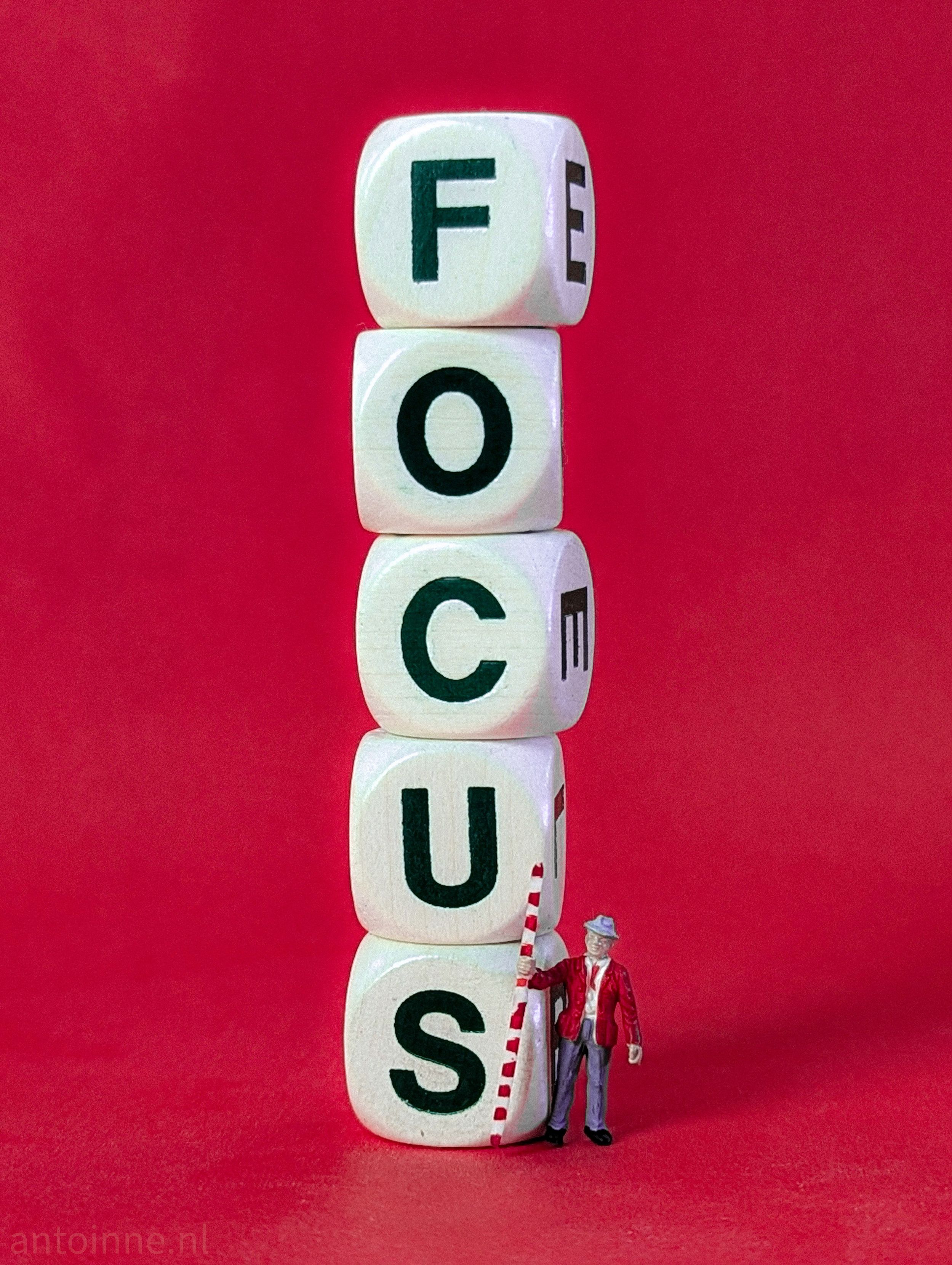 The main subject is a stack of five wooden dice with letters. Each block has a black letter on it, and together, they spell the word "FOCUS" vertically. The blocks are slightly off-kilter, giving the stack a sense of fragility or precariousness.

At the base of the stack, there is a small miniature figurine of a man. He is dressed in a red jacket and a hat, and he is holding a red and white striped pole, which he appears to be using to measure the stack of blocks.