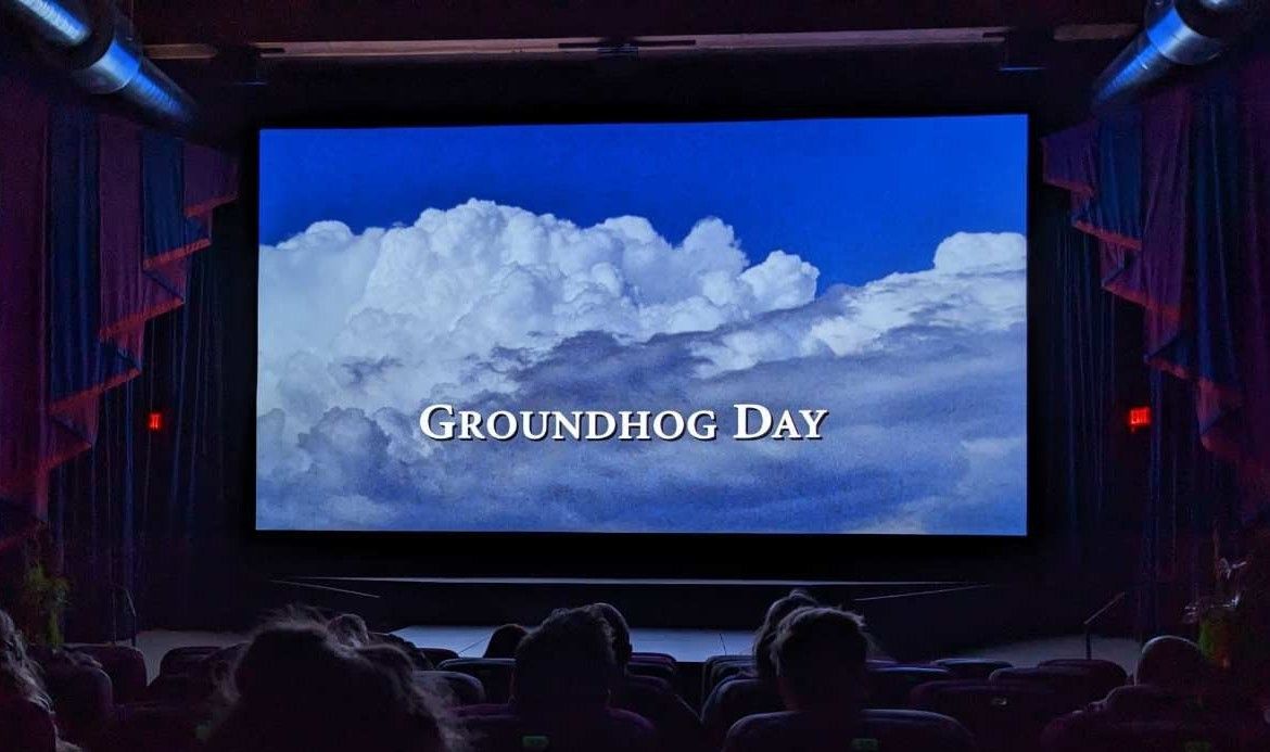 A theater screen with a bright blue sky and cloudy day. Text over the clouds reads Groundhog Day.