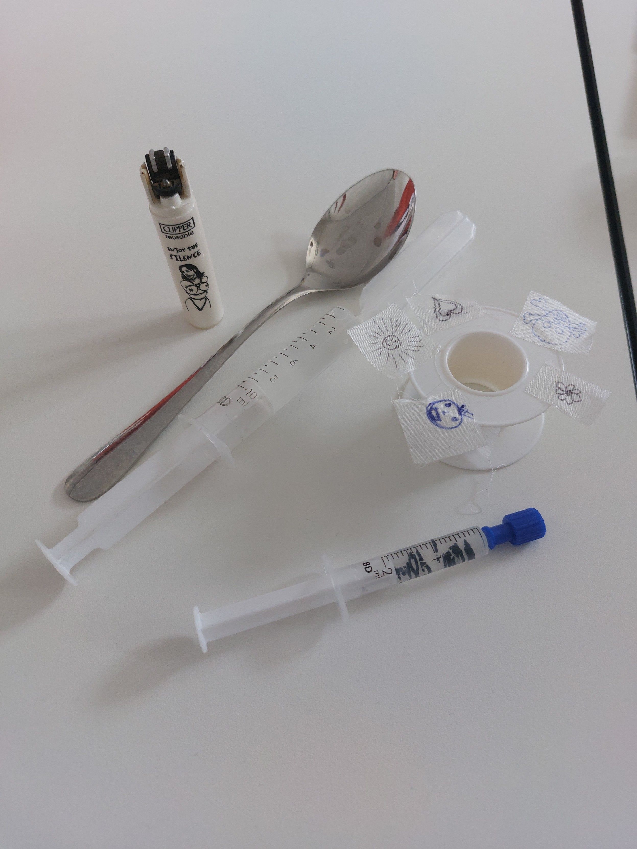 Stuff on a white table: Two filled syringes, a roll of medical tape with pieces of tape on it that have a smiley face, a heart, a sun, etc. on them, making them look like edibles, a spoon, and a lighter with a BDSM drawing on it. It looks like something you would find in the backrooms of a shady club.