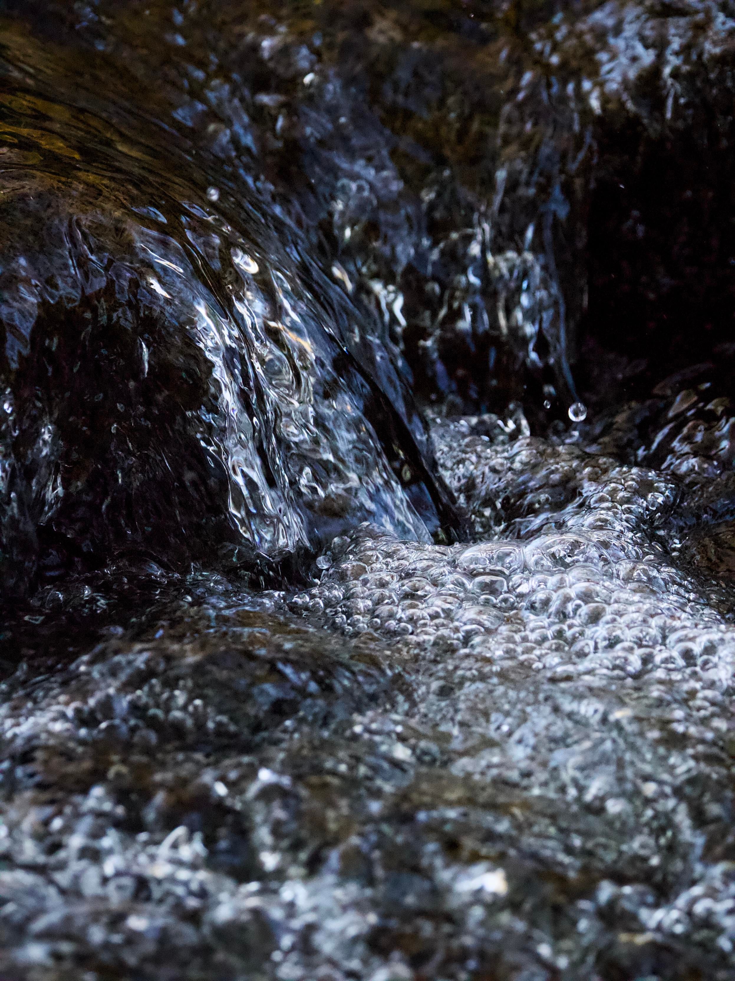 Water pouring over rocks in a stream is captured with a very fast shutter speed to freeze the moment when bright sunlight pierces the trees to highlight the hundreds of tiny bubbles forming on the surface of the water