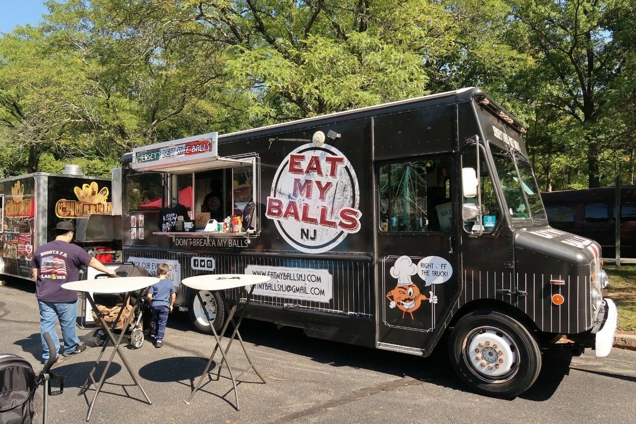 A black panel food truck, with a little cafe set up on the side, with a round white ad red logo on the side which says EAT MY BALLS.