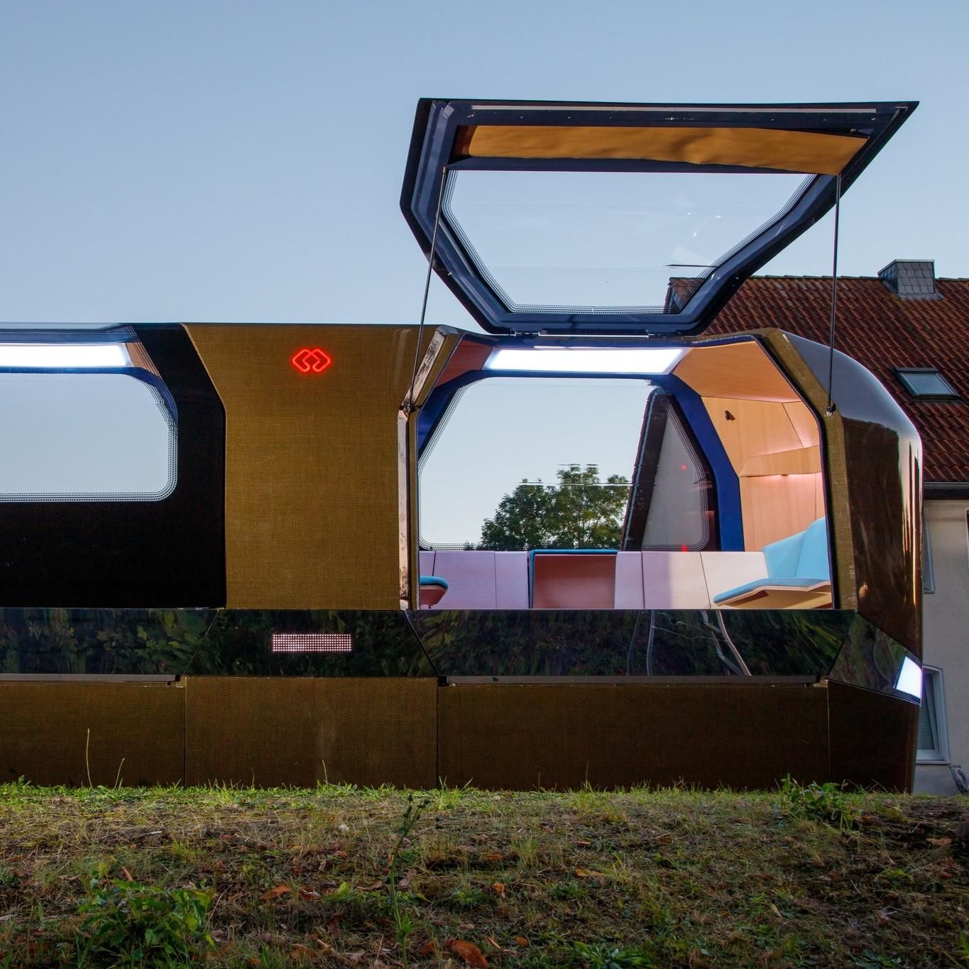 This image shows a gyro-stabilized monorail cabin. The cabin has a reflective, metallic exterior with large windows on the sides with one of them being open and revealing a cozy interior with seating and wood accents. It is situated outdoors on a patch of grass, with residential houses in the background and a clear sky overhead. 
