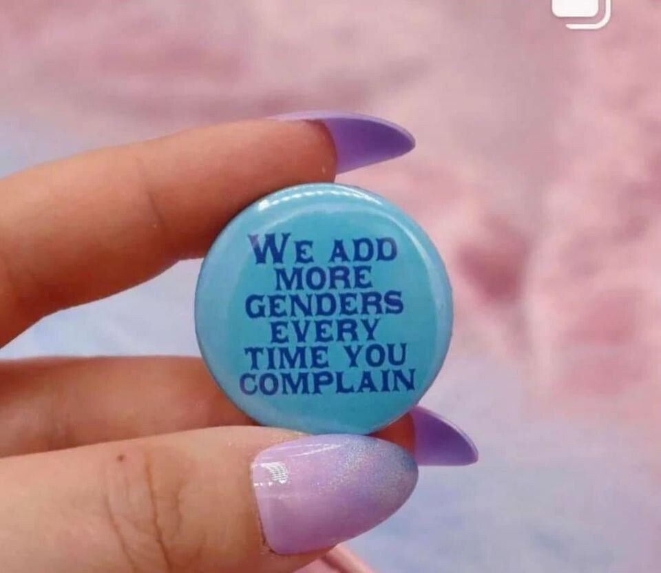 A hand with long violet nails holds a button reading "We add more genders every time you complain"